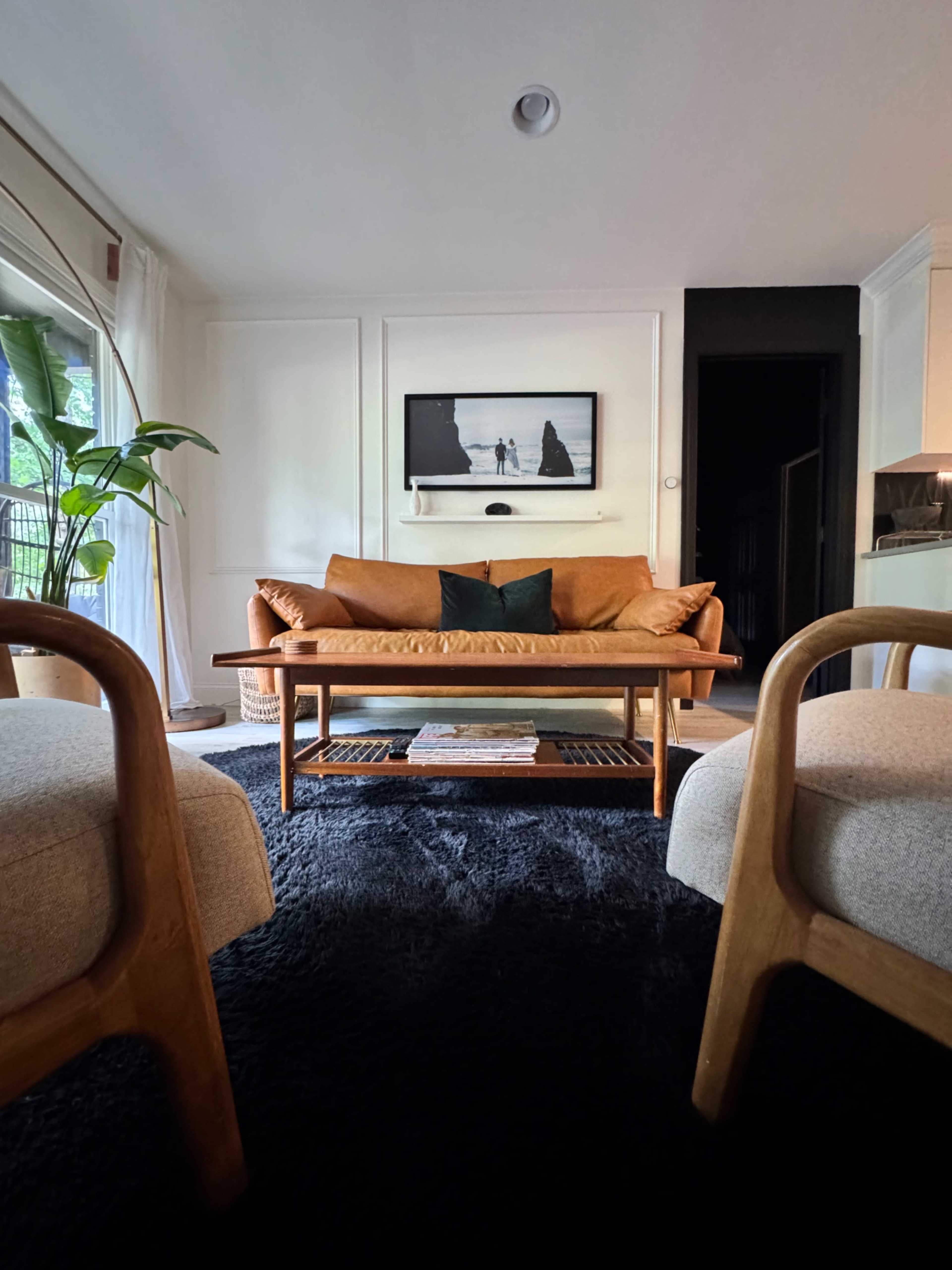 The image shows a cozy living room with a brown leather sofa, a wooden coffee table, and two light-colored armchairs, all set on a dark rug.