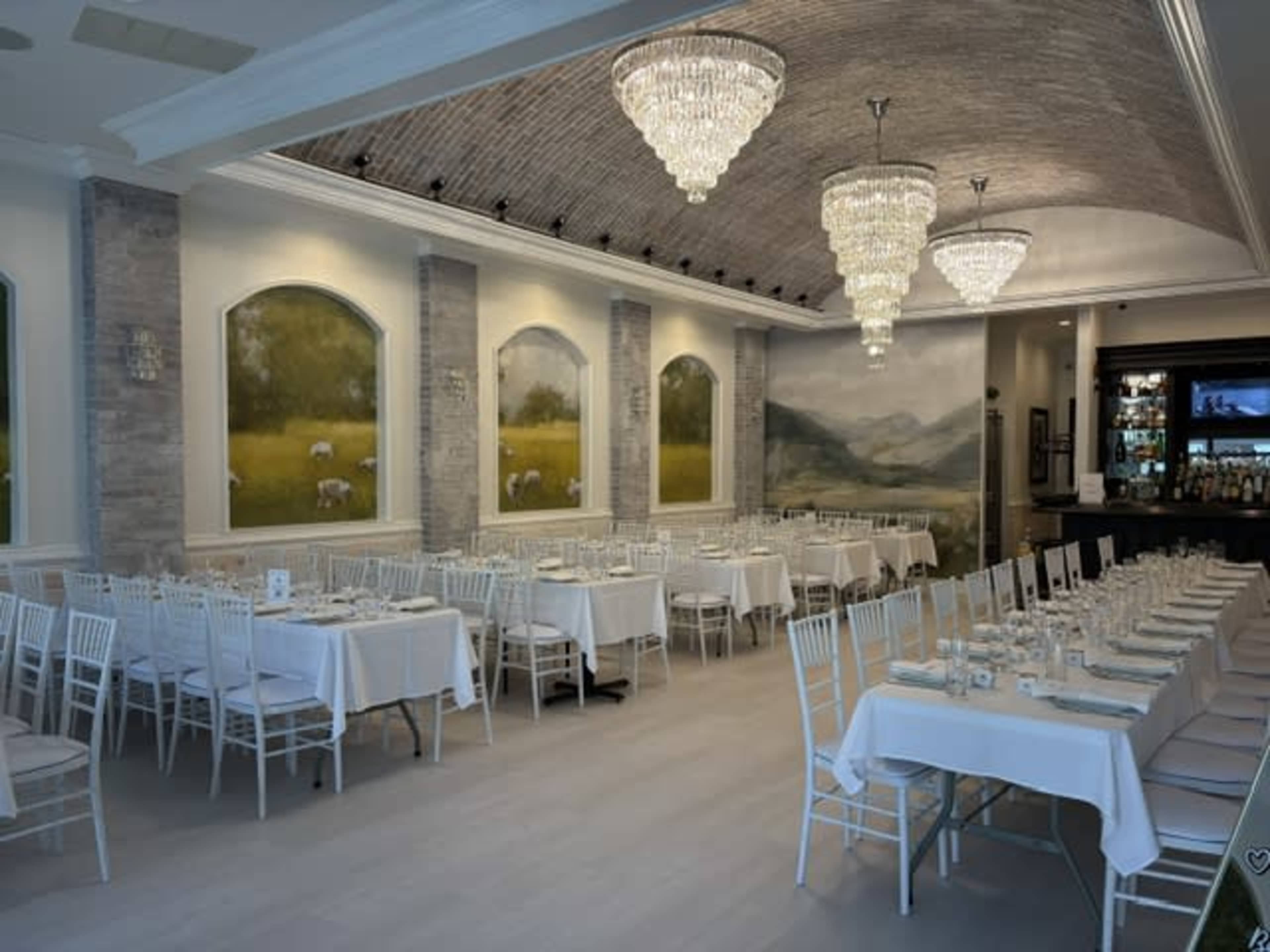 The image shows a spacious restaurant interior featuring white tablecloths, rows of empty tables with chiavari chairs, and decorative chandeliers overhead.