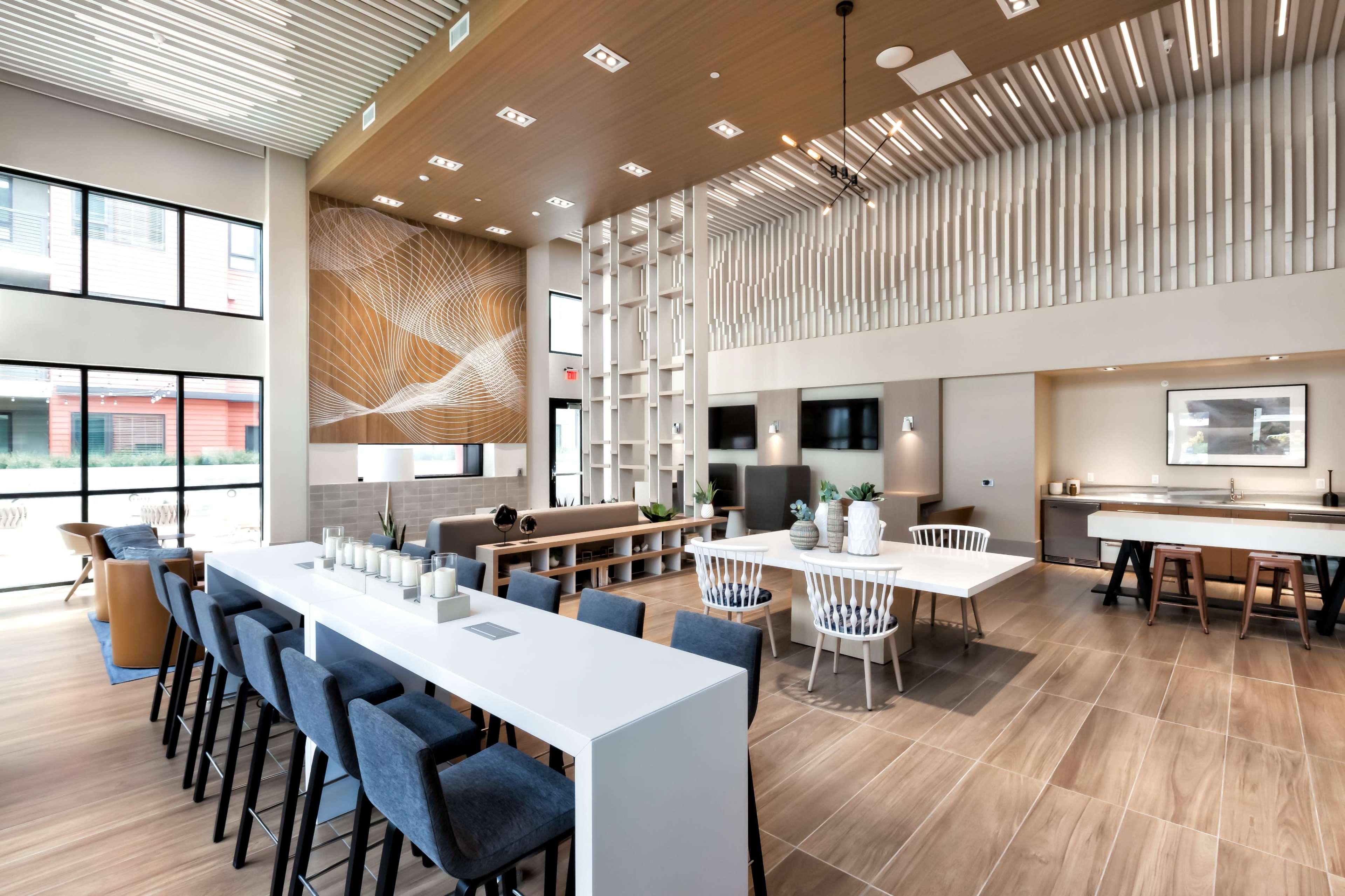 An interior communal lounge area with modern furniture, large windows, and a mix of seating arrangements around a long table and a bar-height counter.