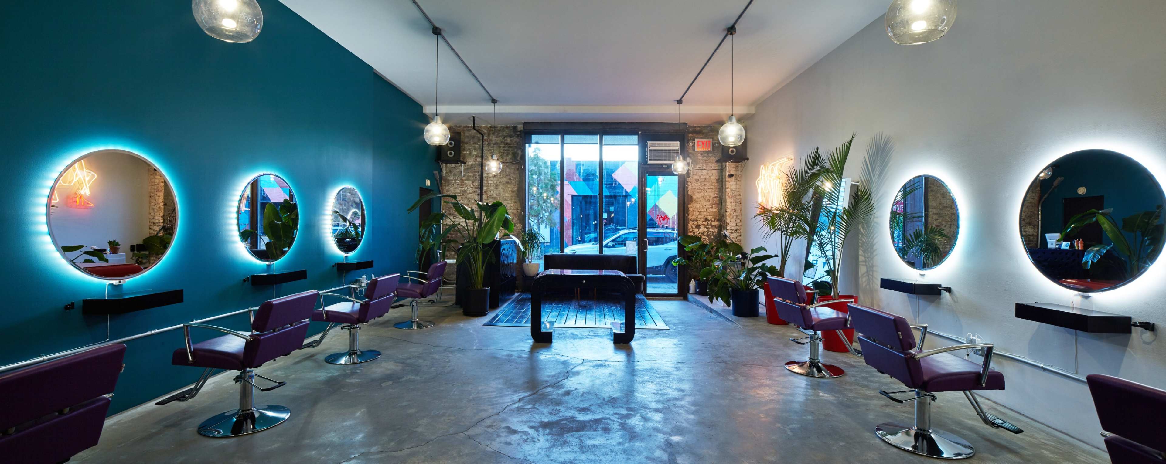 A modern hair salon with circular mirrors and chairs arranged around the space, featuring plants and large windows that allow natural light to enter.