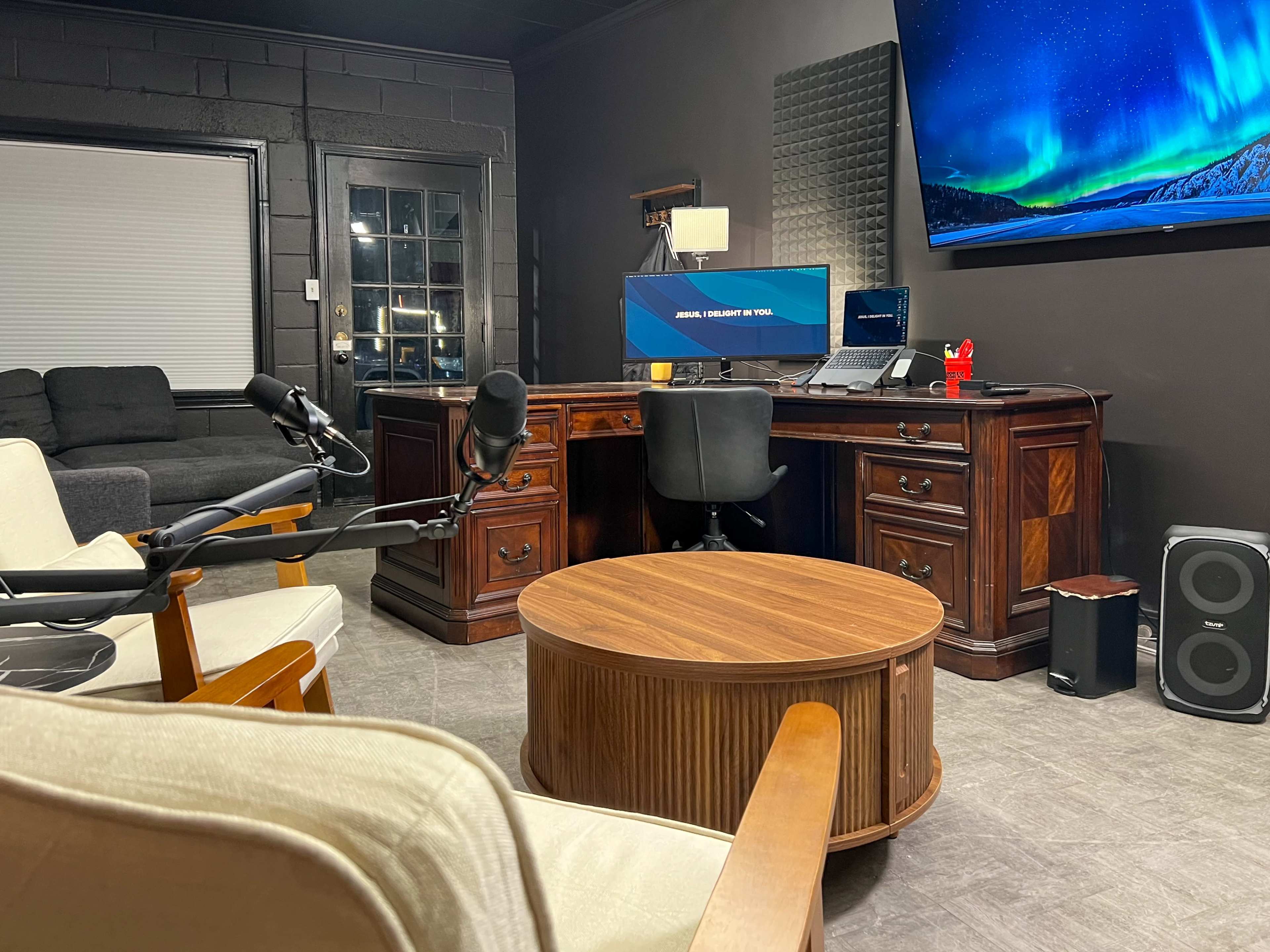The image shows a modern office space featuring a large wooden desk, a black chair, and a round coffee table, with a couch and a flat-screen monitor displaying a scenic background.