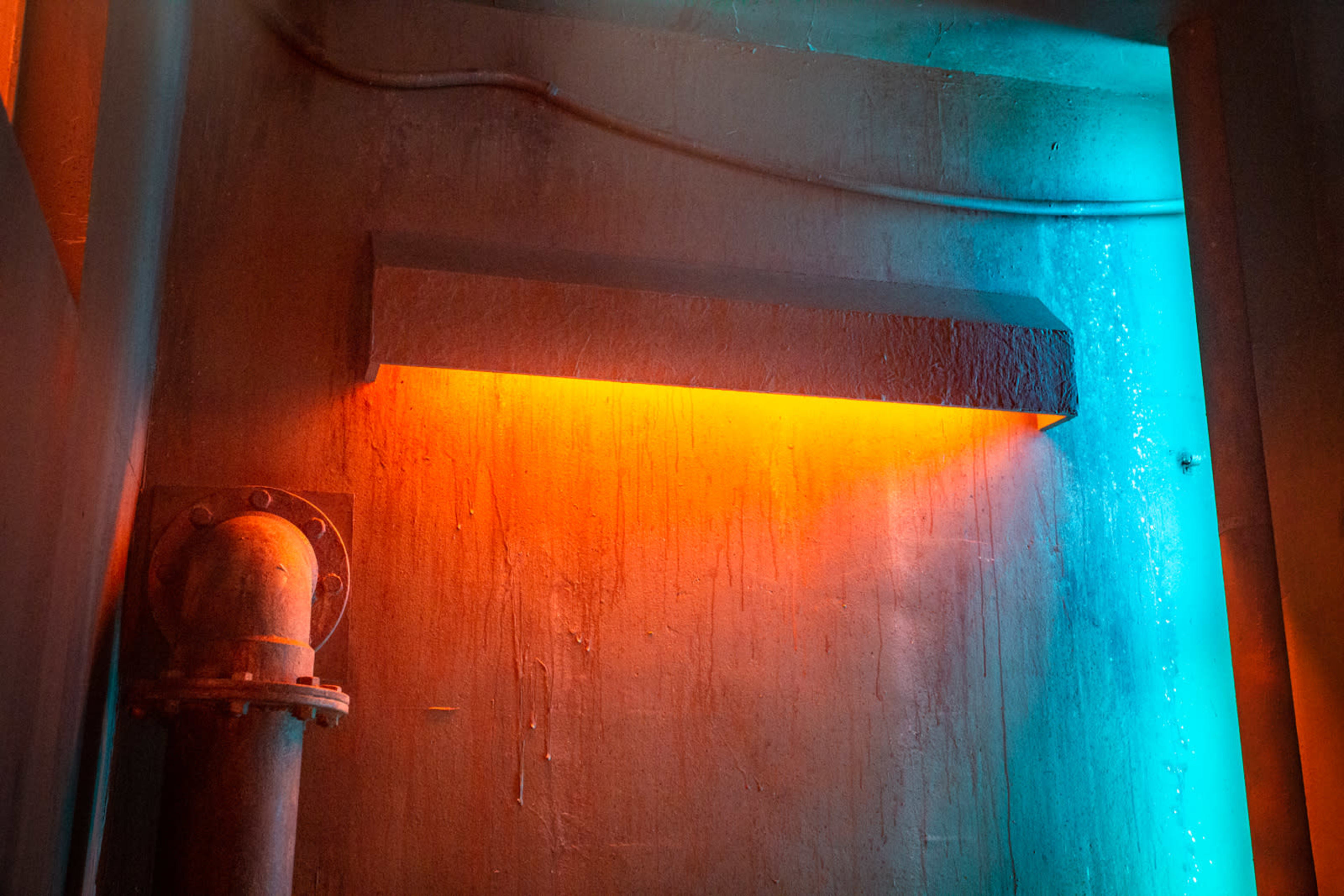 A glowing orange light fixture is mounted on a textured wall beside a large metallic pipe.