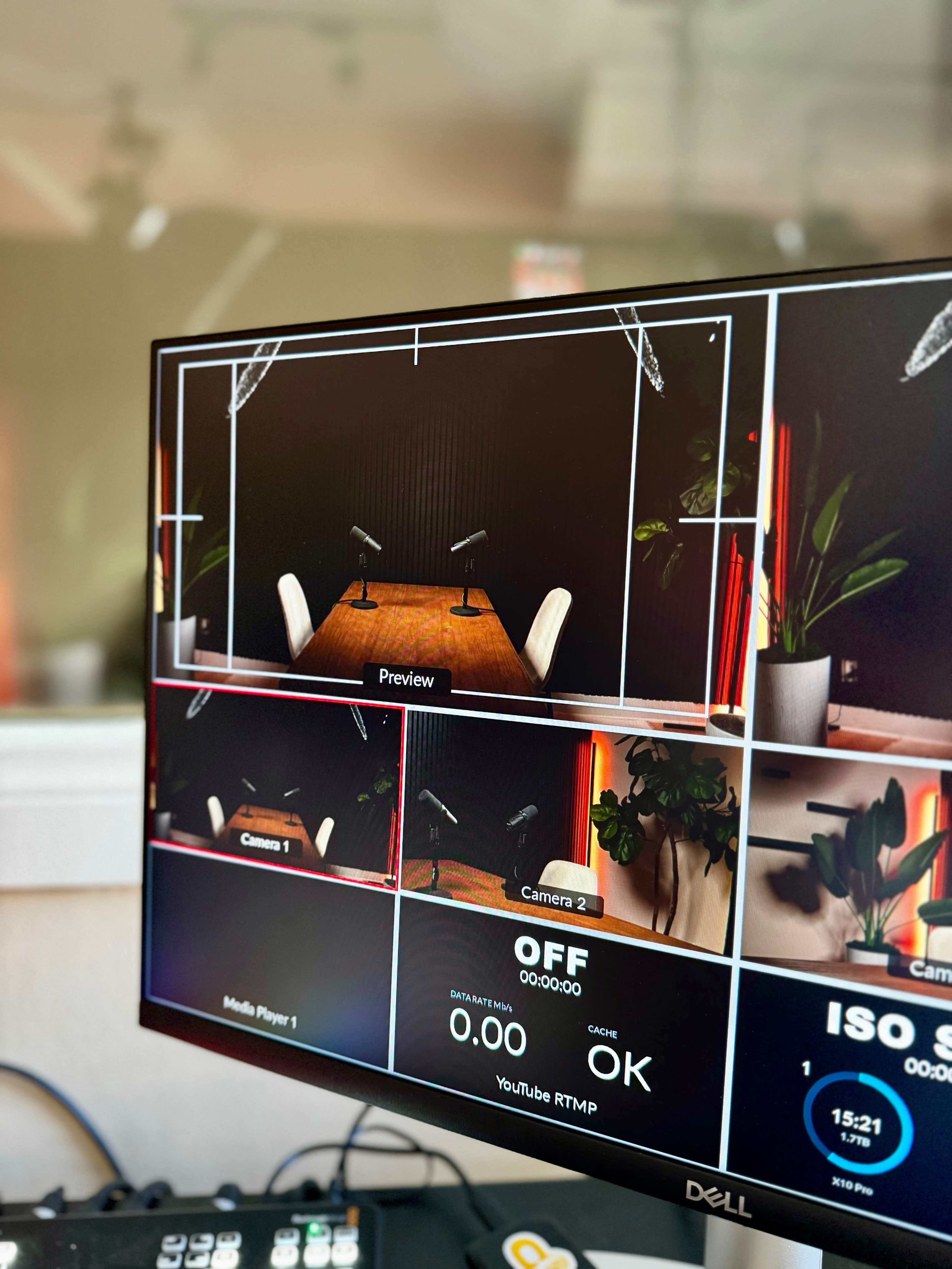 A computer monitor displaying a multi-camera preview setup for a video production, featuring a table with two microphones and plants in the background.