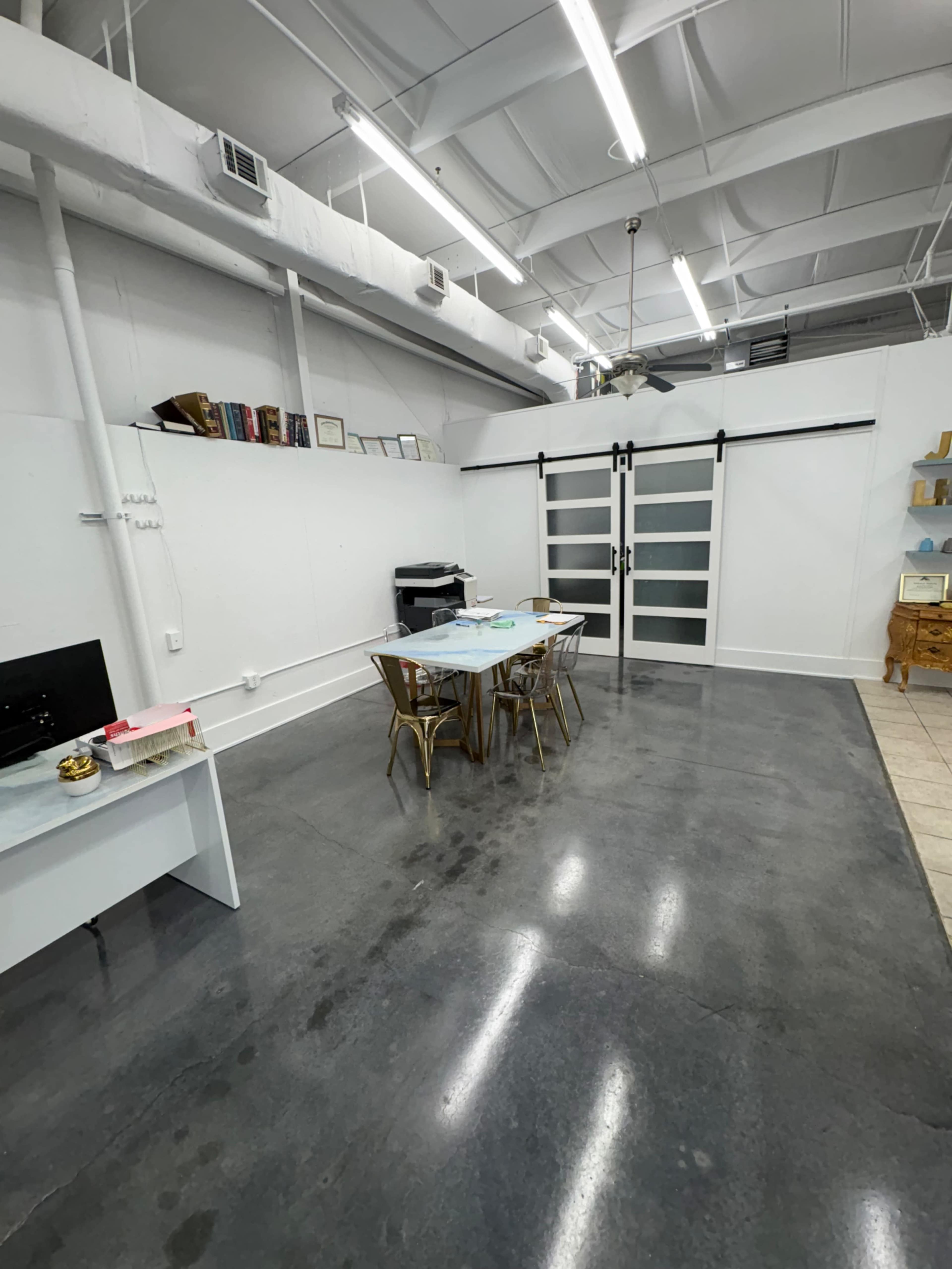 The image shows a spacious, minimally furnished room with a table and chairs, a printer, and sliding glass doors, all on a polished concrete floor.