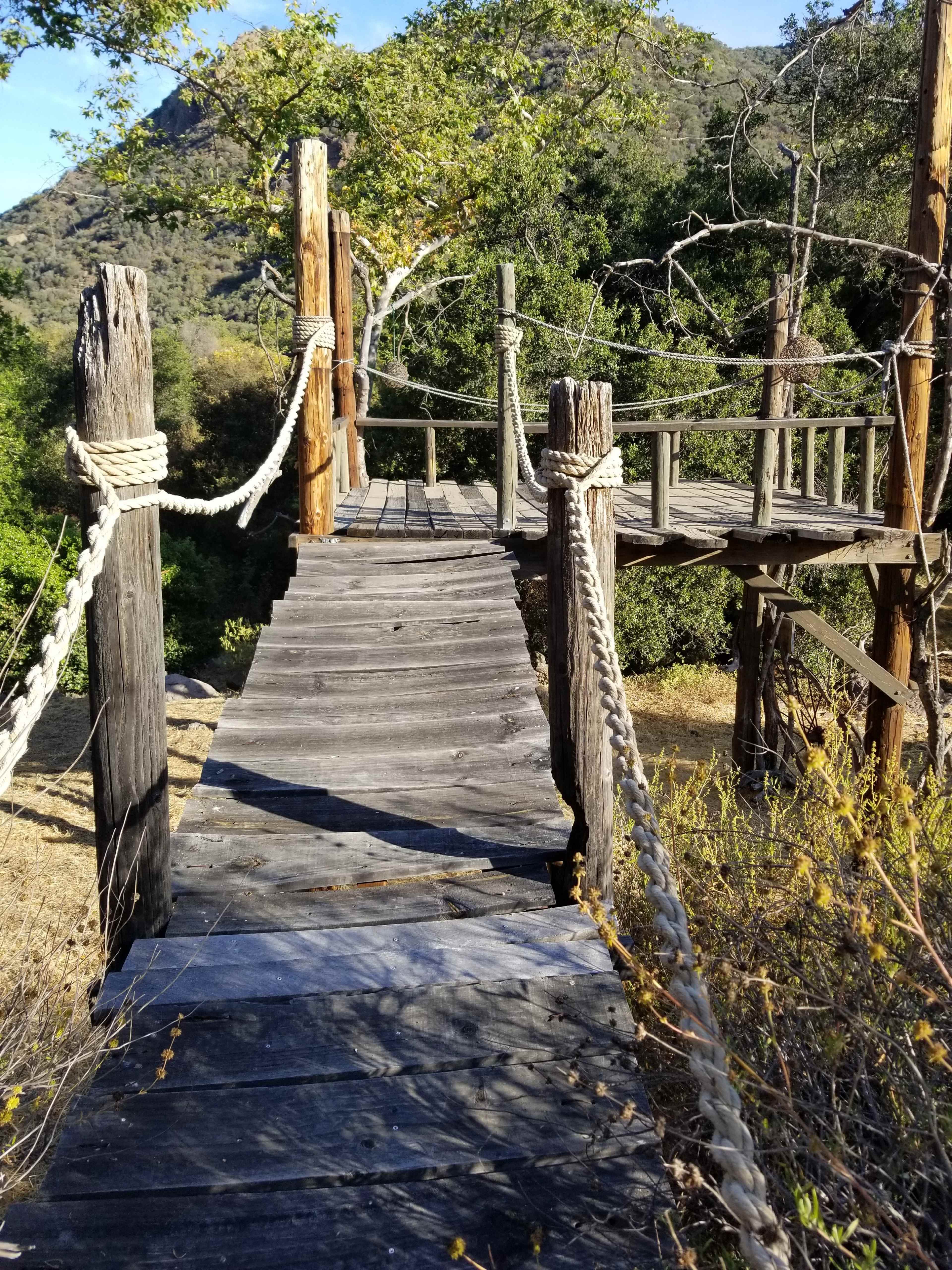 A wooden walkway with rope railings leads to a small elevated platform surrounded by greenery and hills.