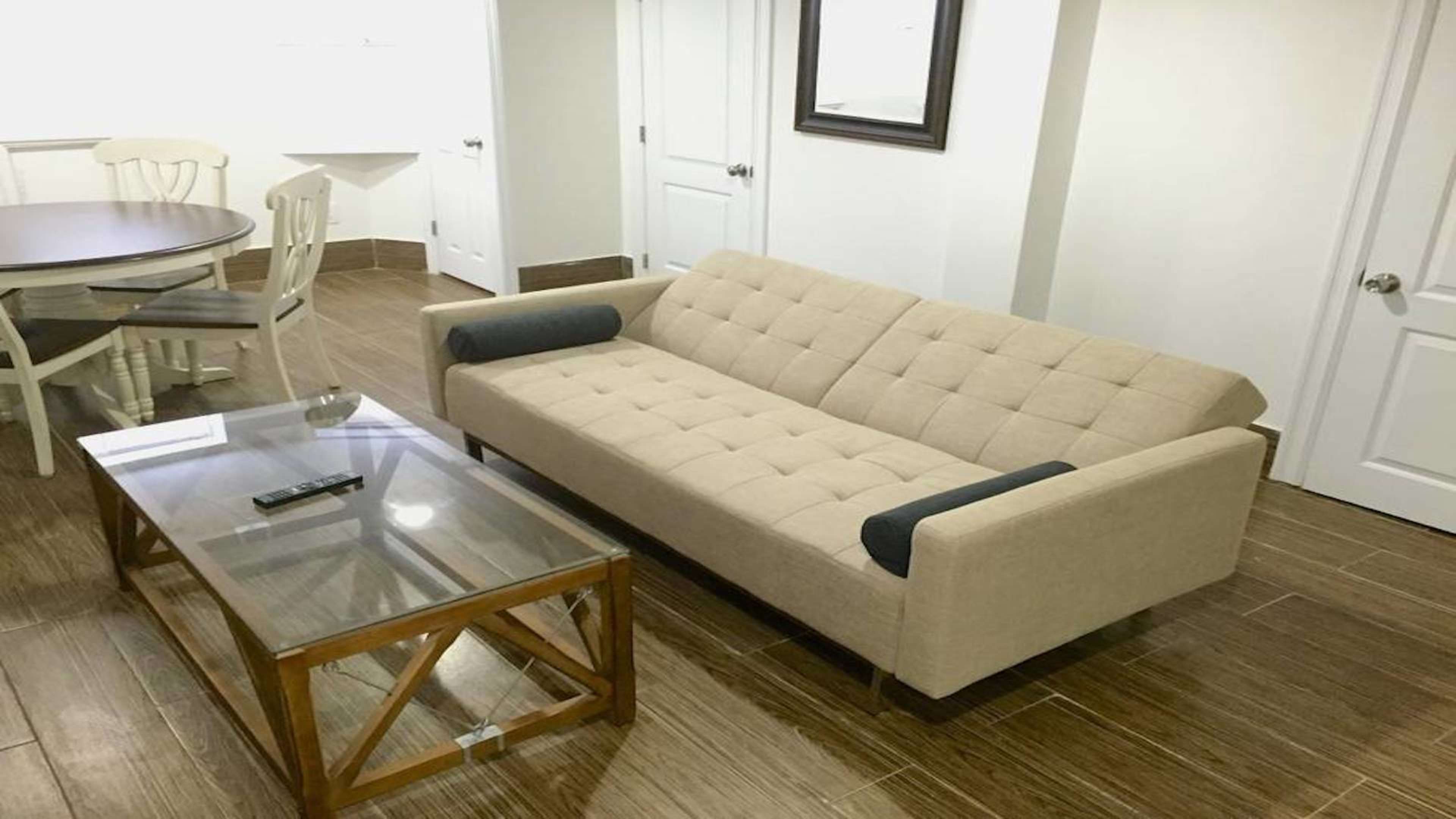 A light-colored sofa with dual arm cushions is placed in a room featuring a glass coffee table and a round dining table with white chairs.