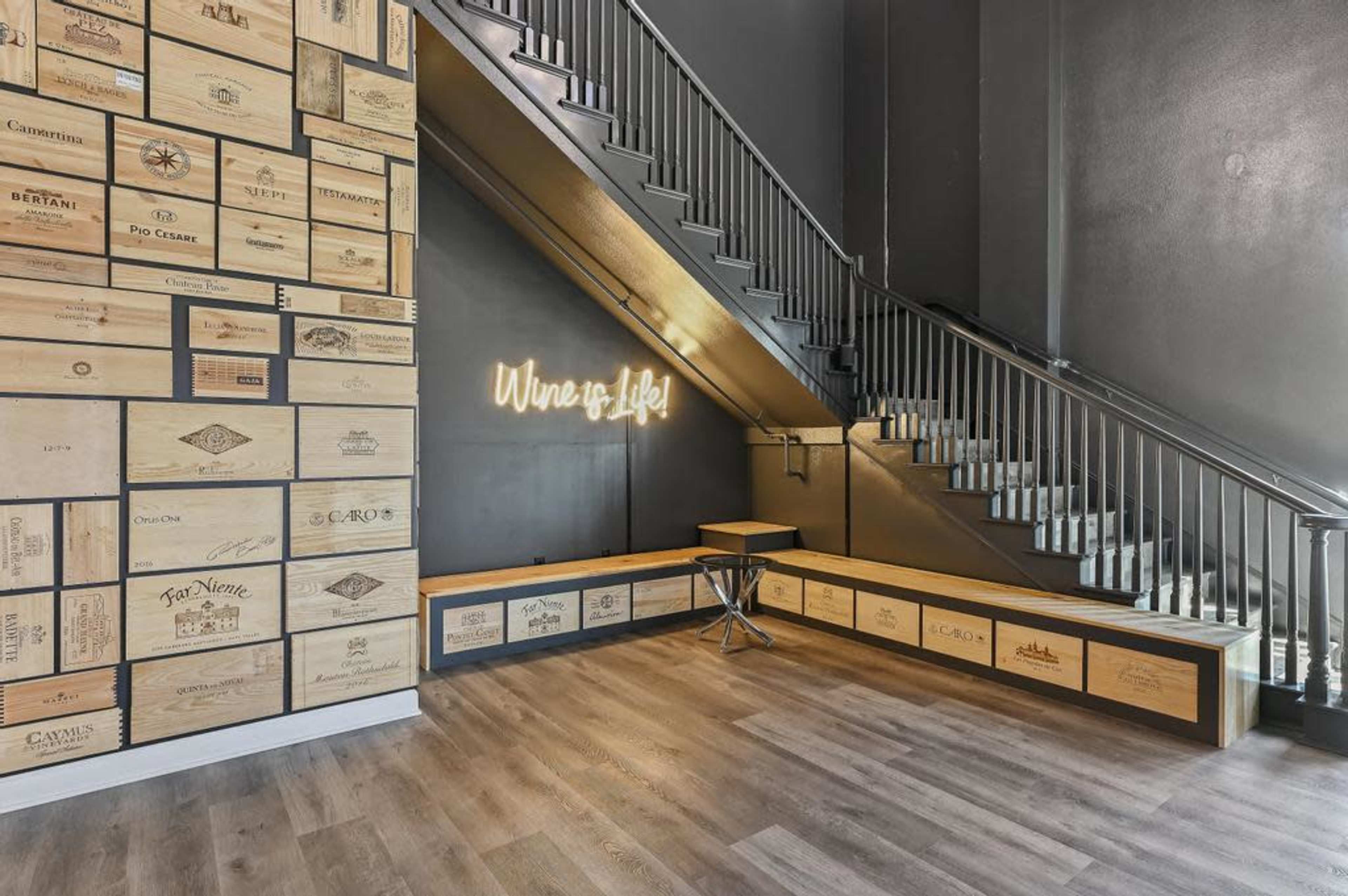 A modern interior space featuring a wooden wall adorned with wine crate labels and a staircase leading to an upper level, with a neon sign that reads "Wine is Life."