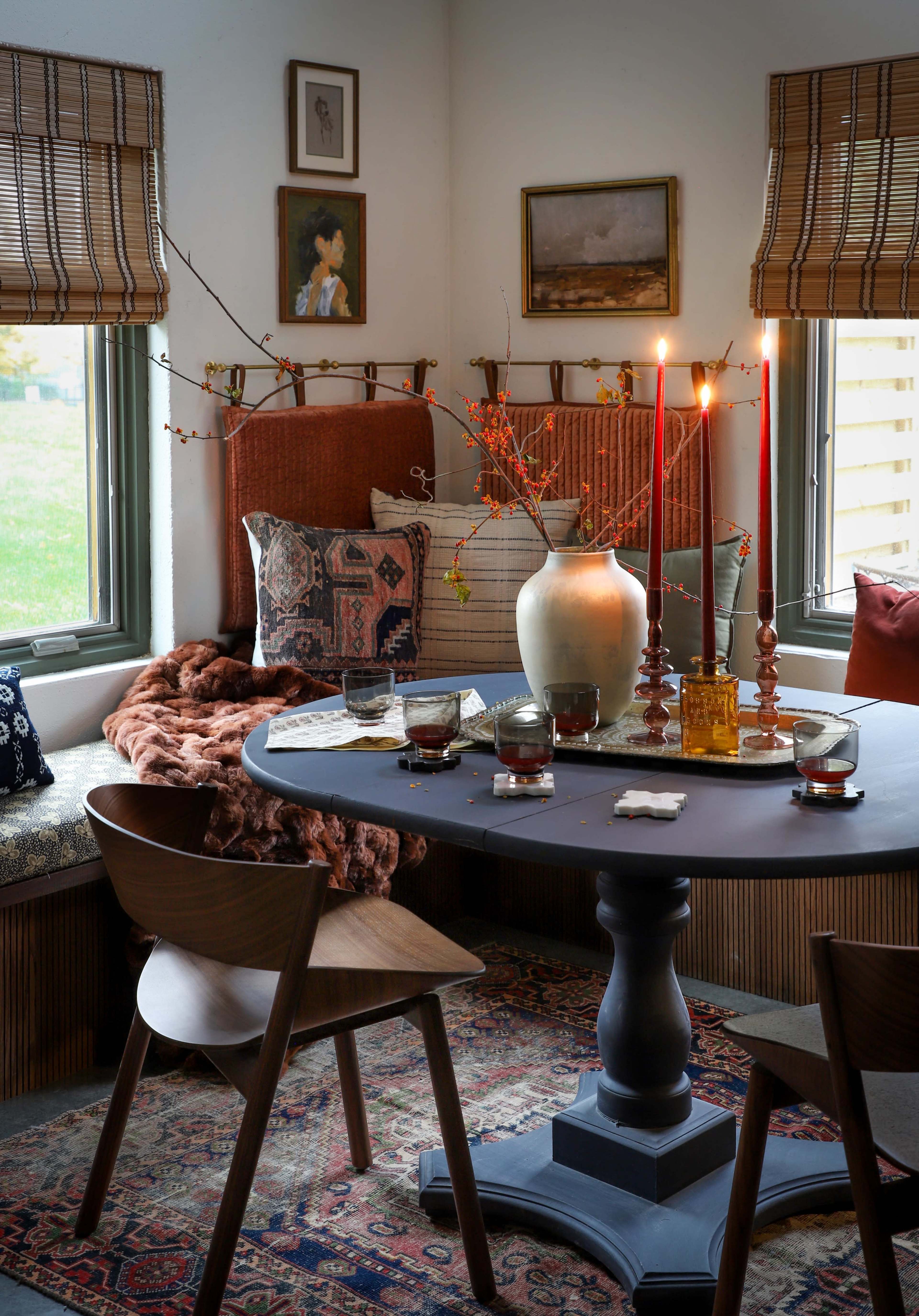 A cozy dining area features a round black table surrounded by wooden chairs, with a vase and candles on the table, while a window seat is adorned with cushions and a throw blanket.
