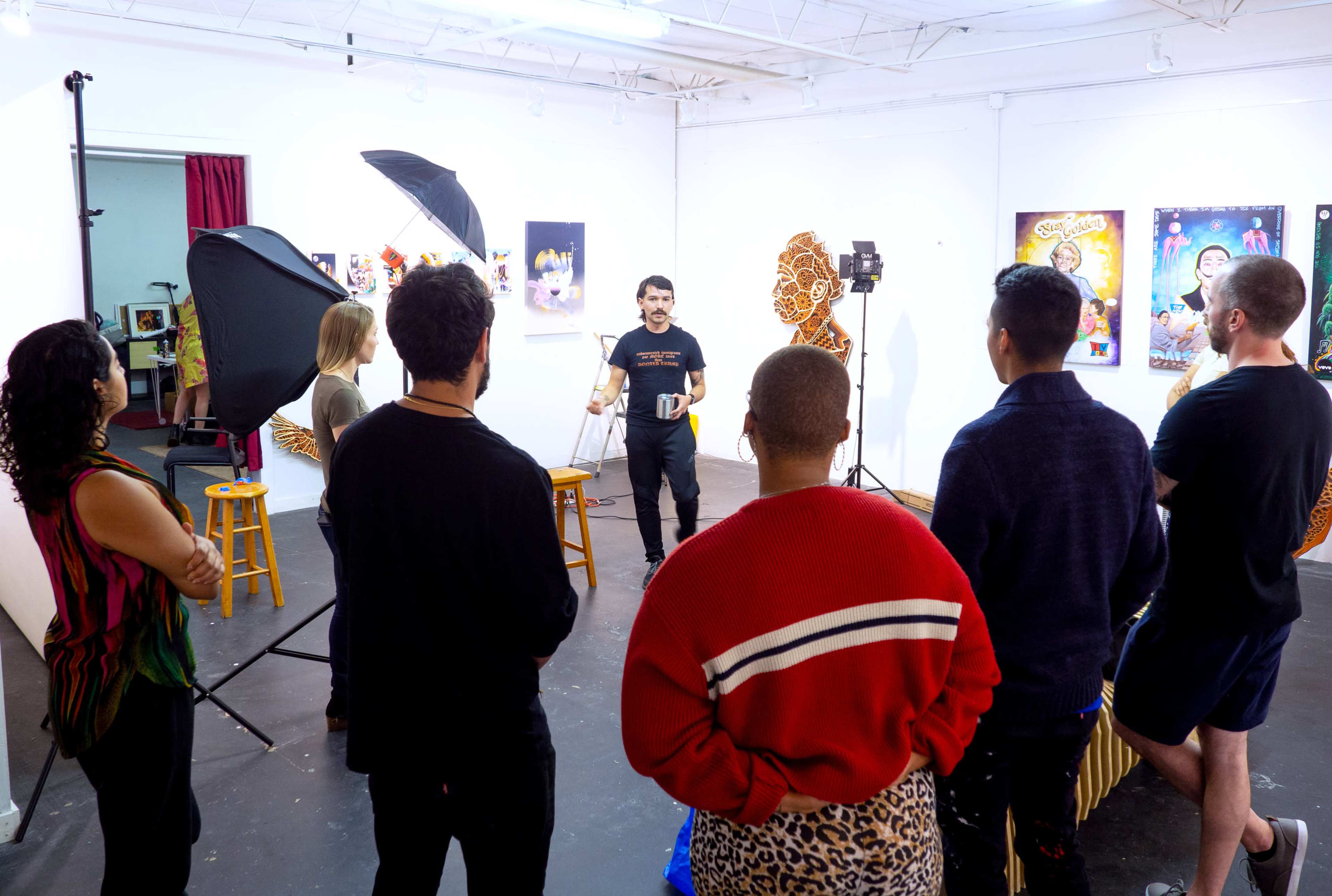 A group of individuals stands in an art studio, facing a person speaking while various artworks are displayed on the walls.