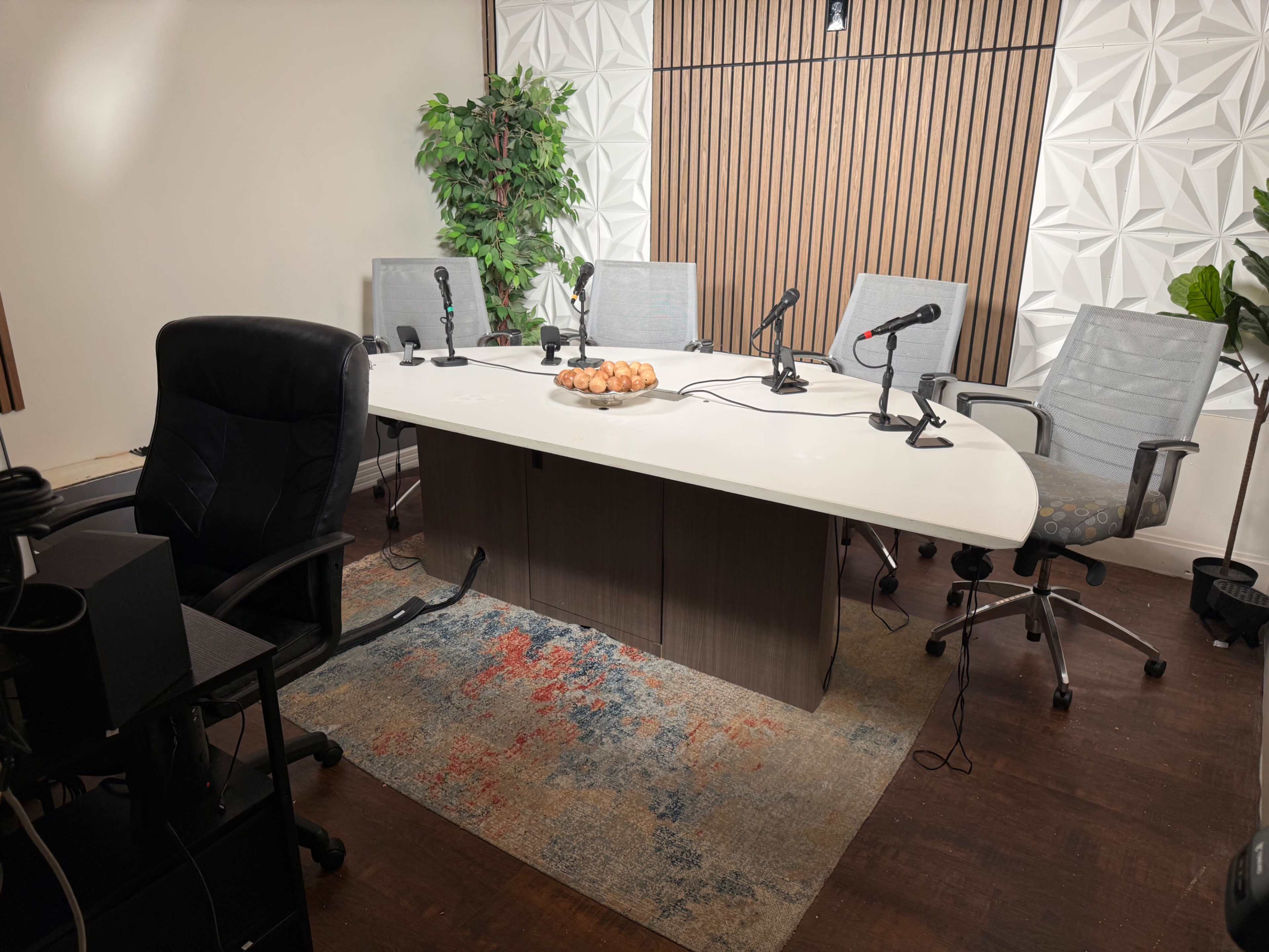 A modern conference room features a large white table surrounded by several chairs, microphones, and a decorative plant, with a bowl of pastries on the table.