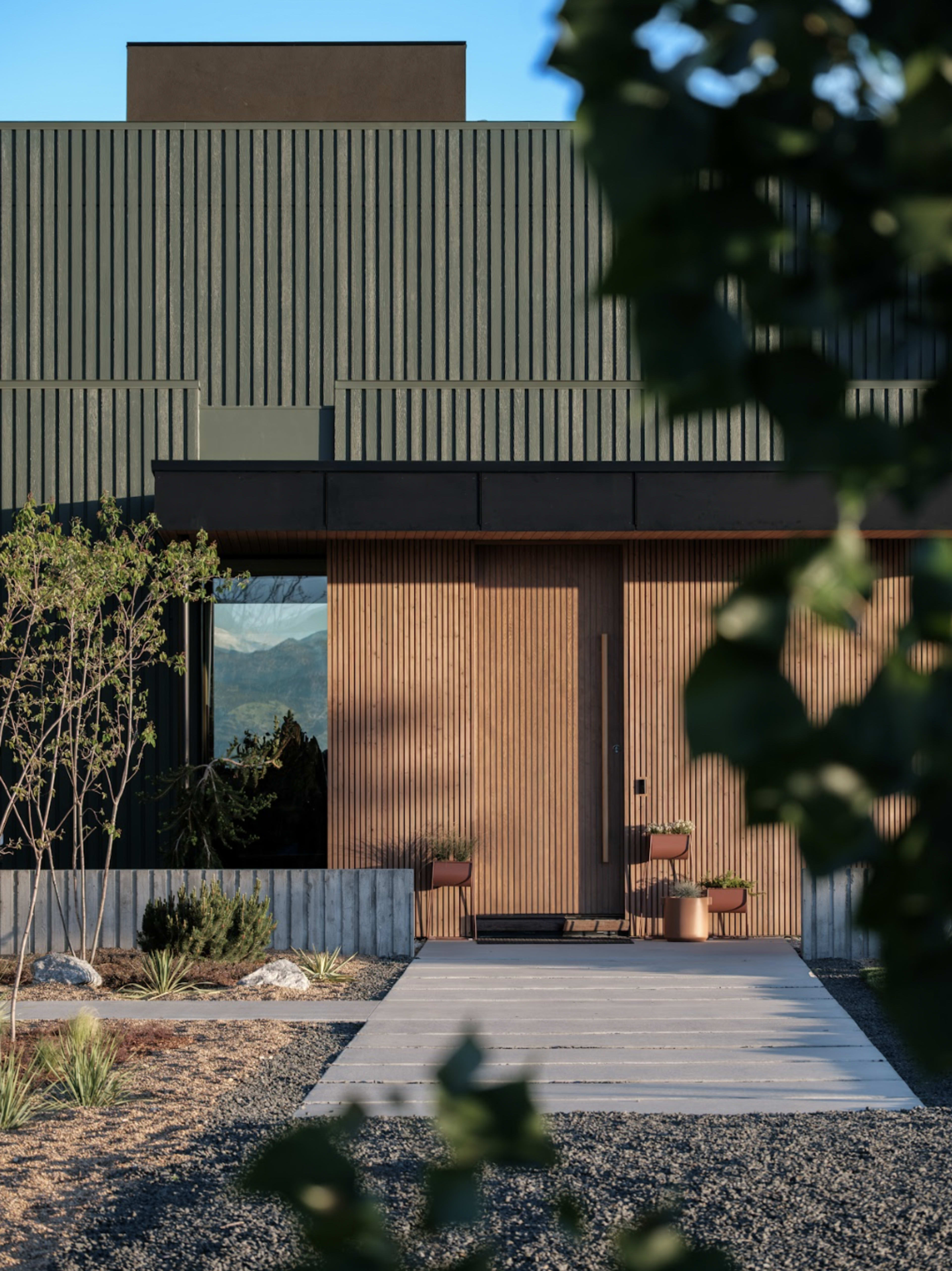 The image depicts a modern house with wooden slats on the exterior and a concrete pathway leading to the entrance, surrounded by minimal landscaping.