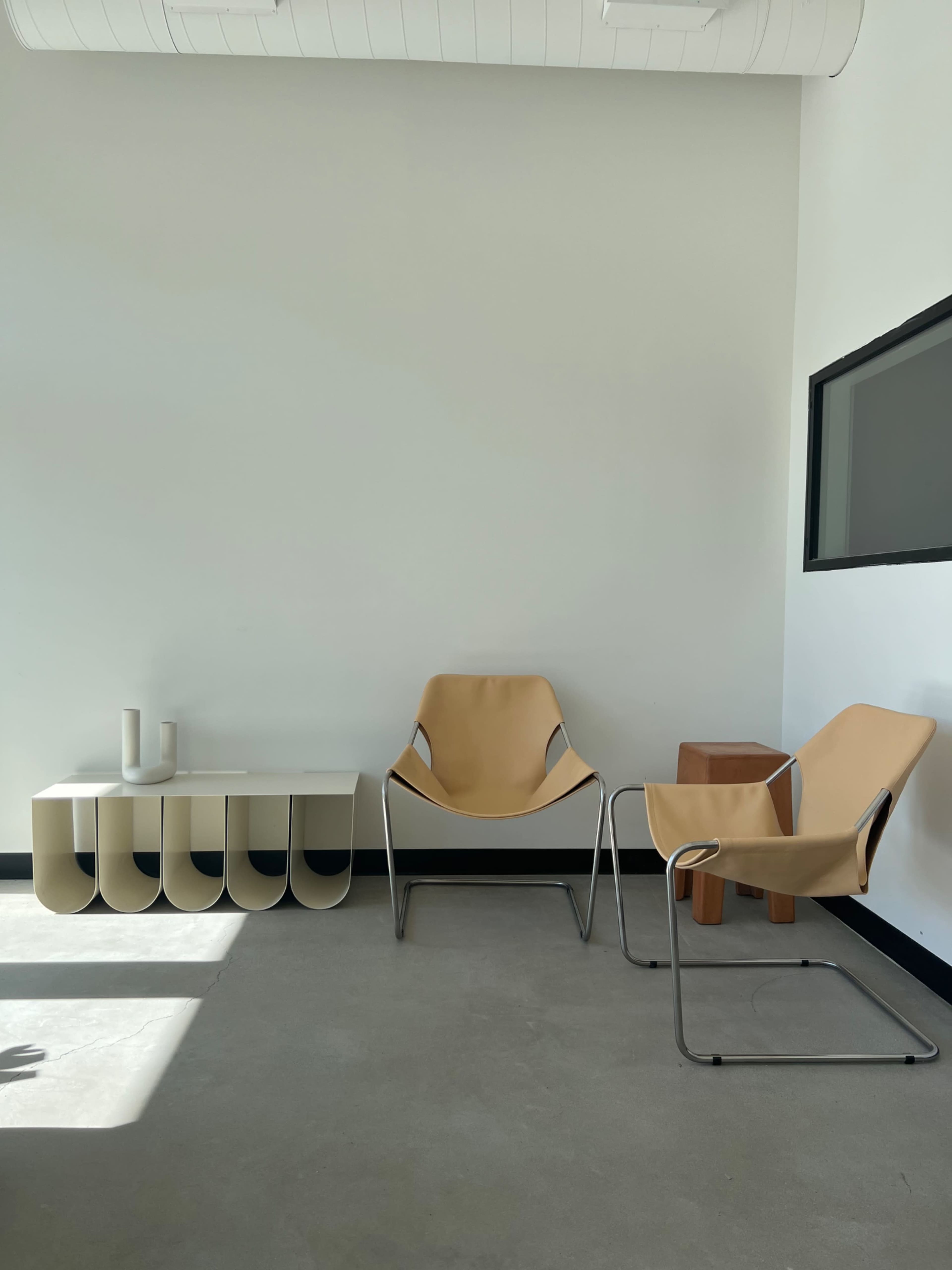 A minimalist interior space featuring two chairs, a small wooden table, and a console with a smooth surface against a plain wall.