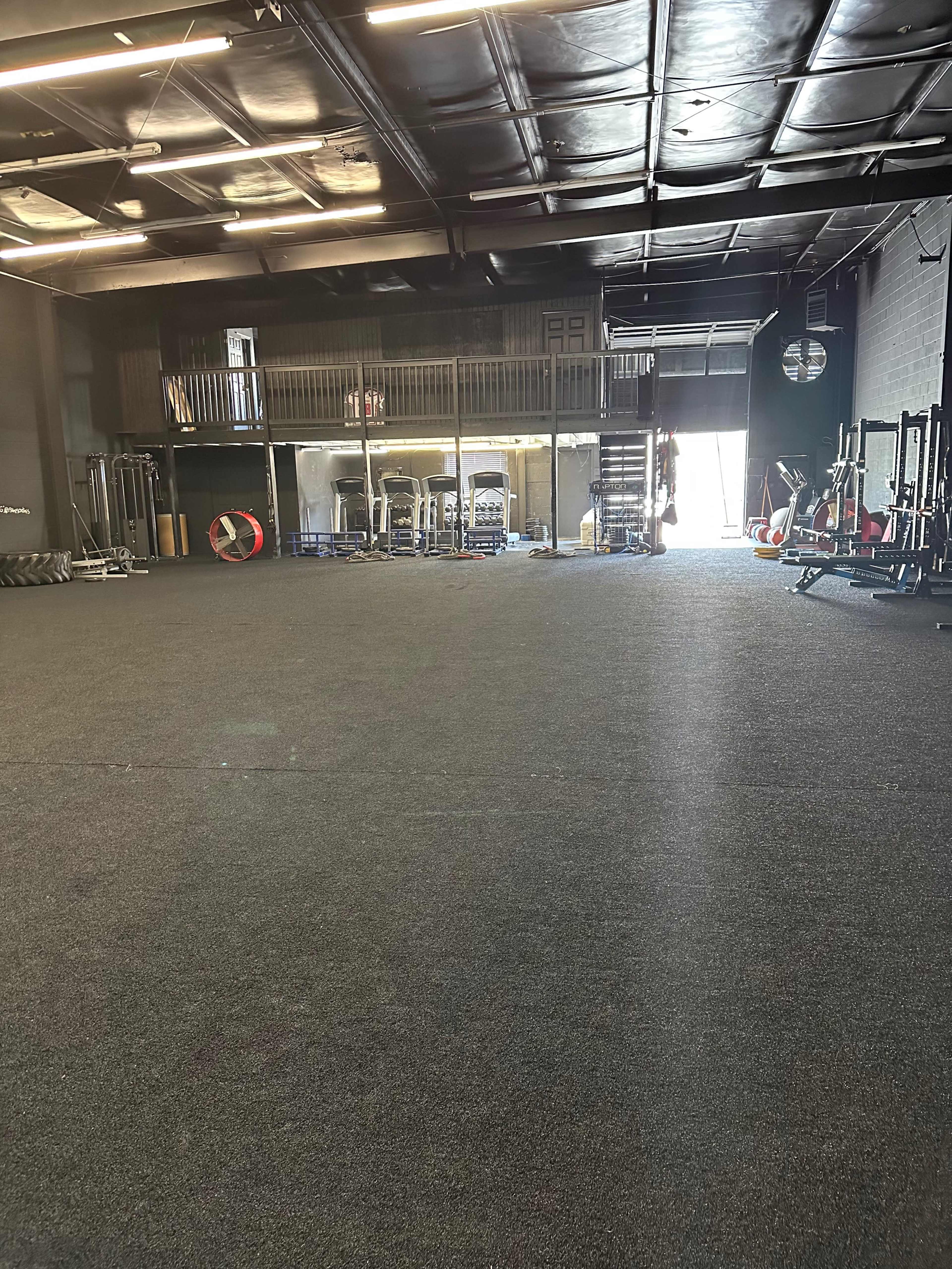 A spacious gym interior with rubber flooring, various fitness equipment, and a mezzanine level in the background.