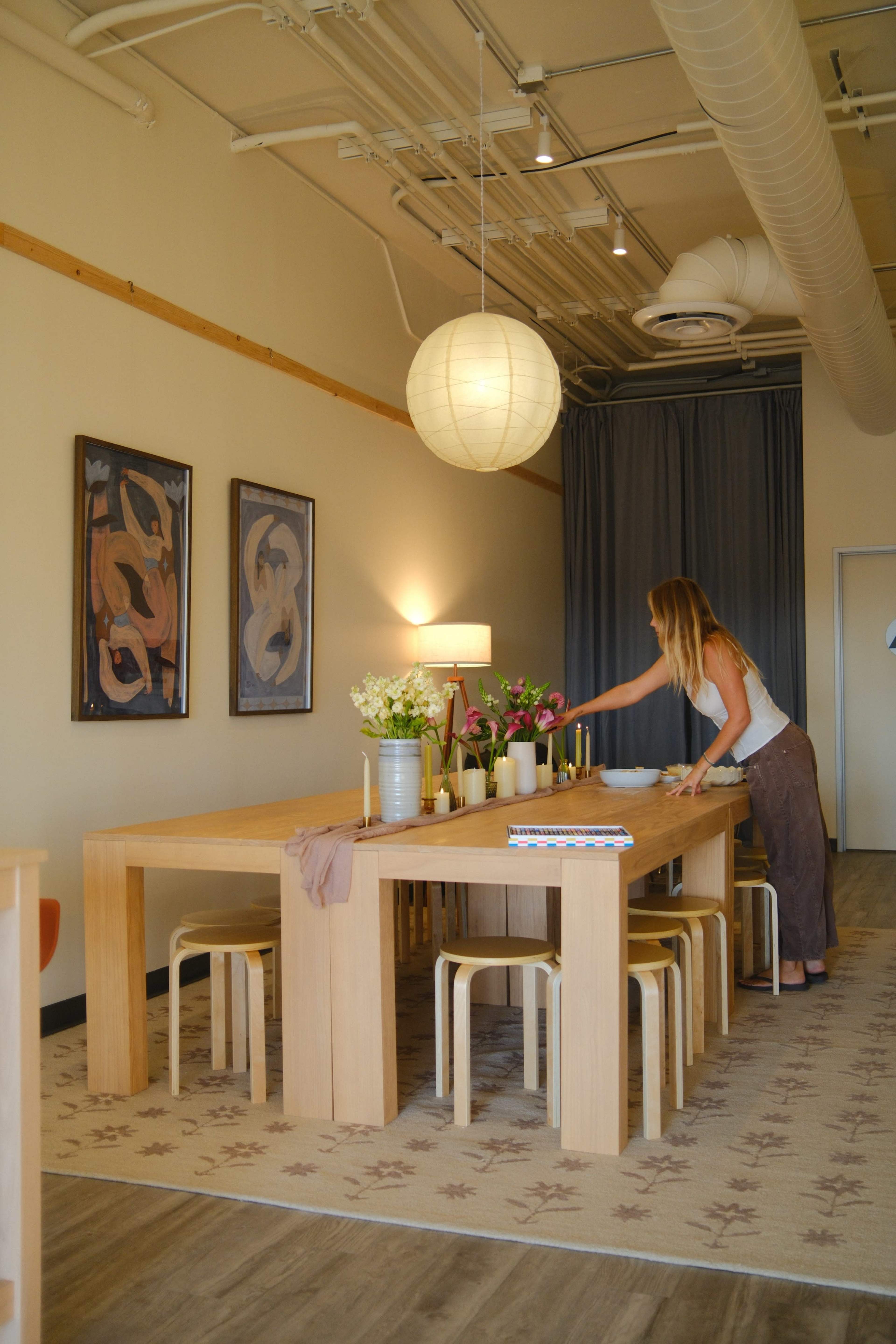 A woman arranges flowers on a large wooden dining table in a softly lit, minimalist room with decorative artwork on the walls.
