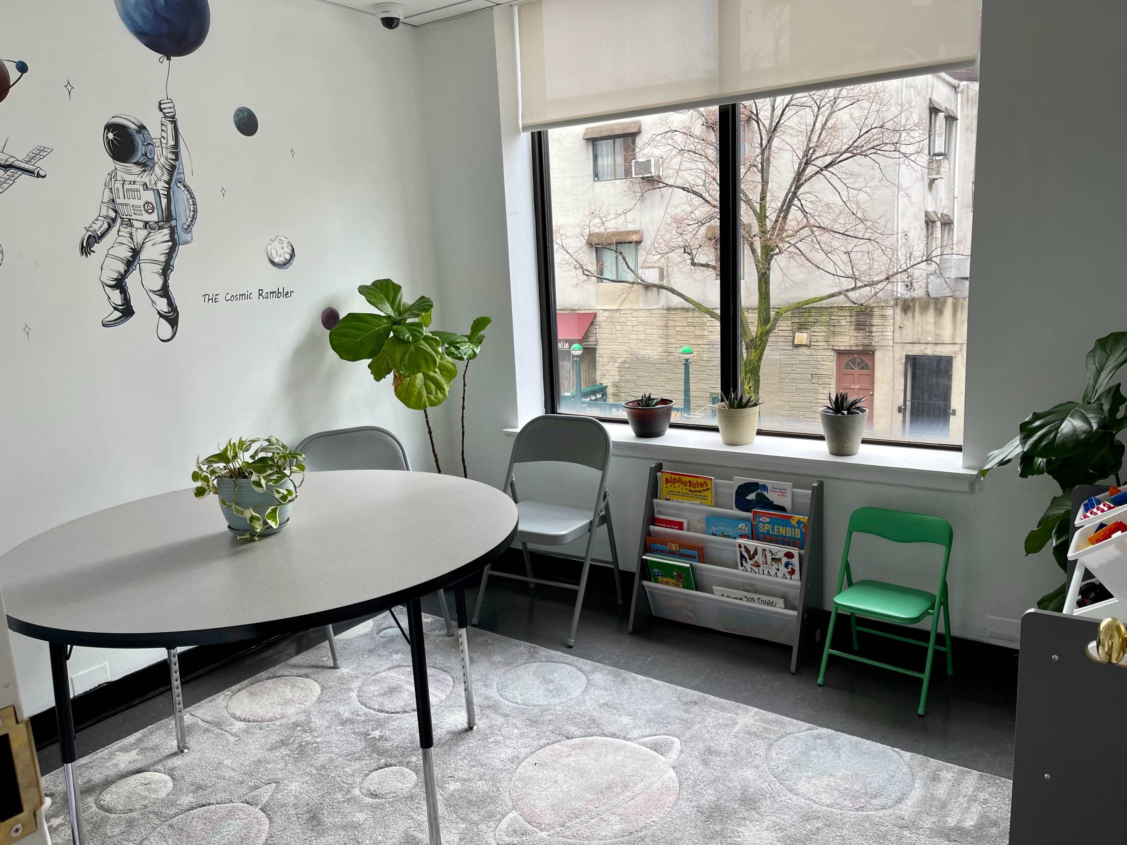 The image shows a small room with a round table, several chairs, a large window overlooking a street, and a wall featuring an astronaut mural.