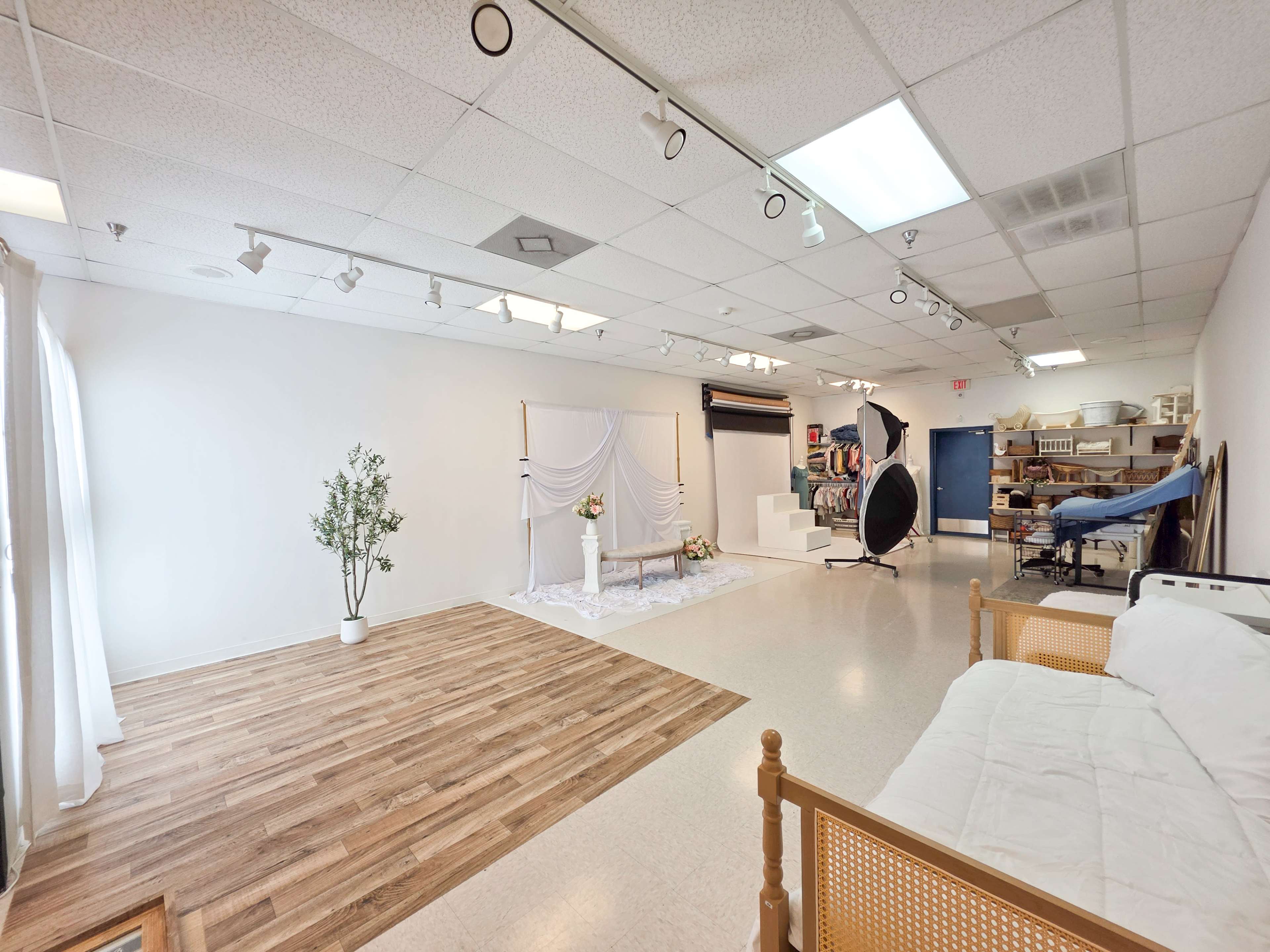 The image shows a spacious photography studio with a wooden floor, white walls, and various props arranged for photo shoots.