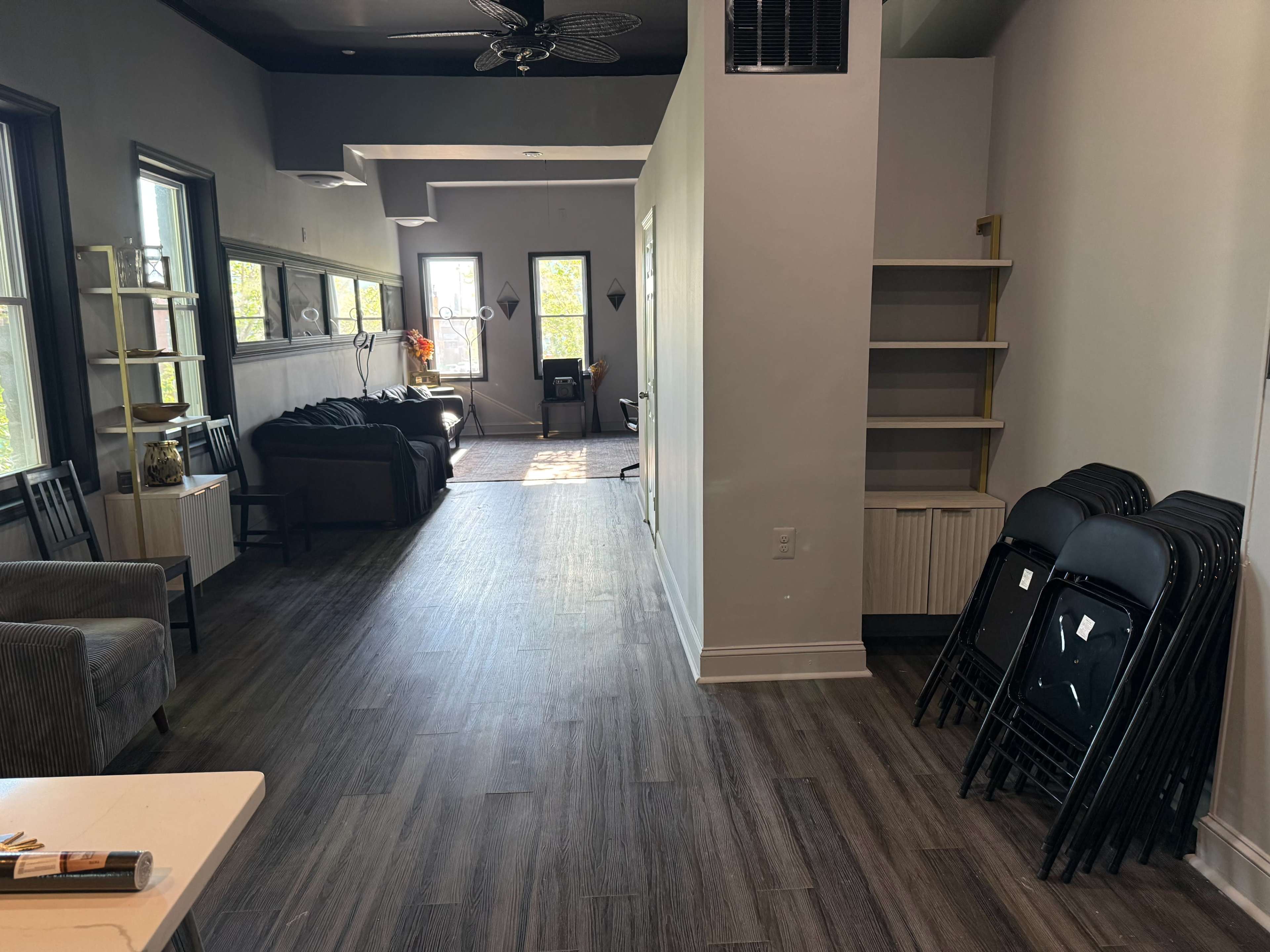 The image shows a spacious interior with a long hallway leading to a living area, featuring a couch, shelves, and several stacked folding chairs on the right.