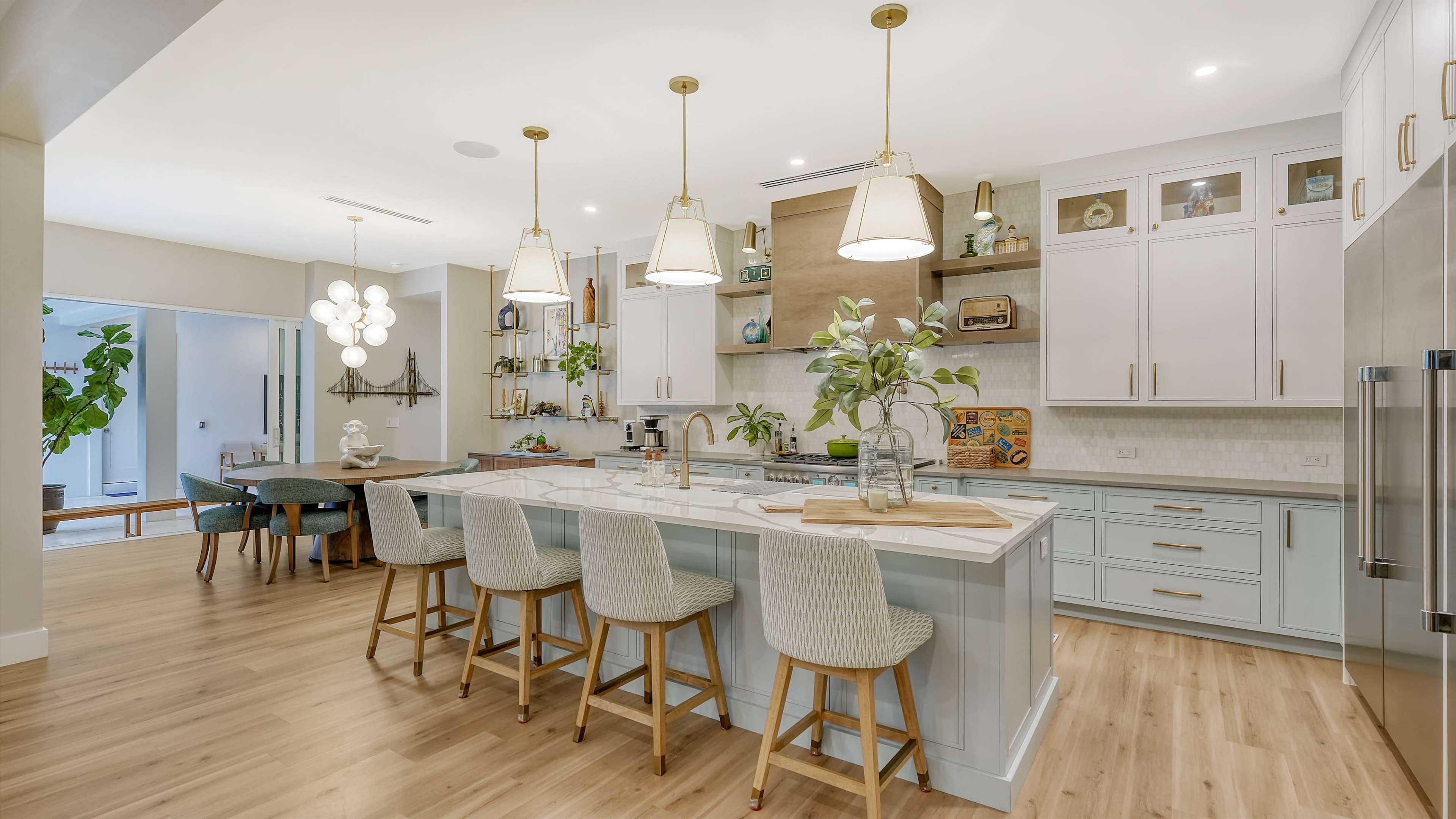 A modern kitchen features a large central island with bar stools, light wood flooring, and various plants and decor elements.