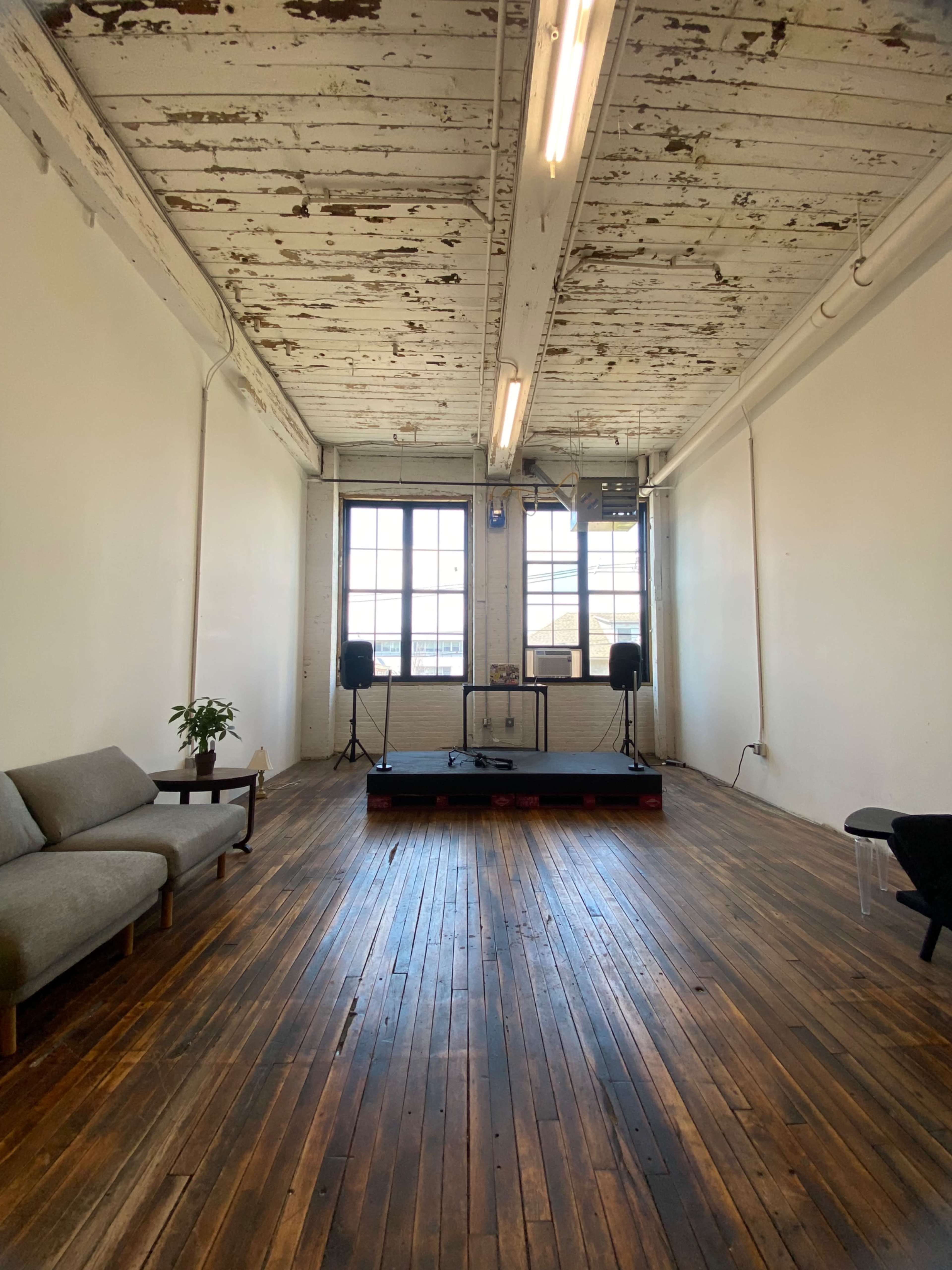 The image shows a spacious room with wooden floors, exposed rafters, large windows, a small stage, and seating.