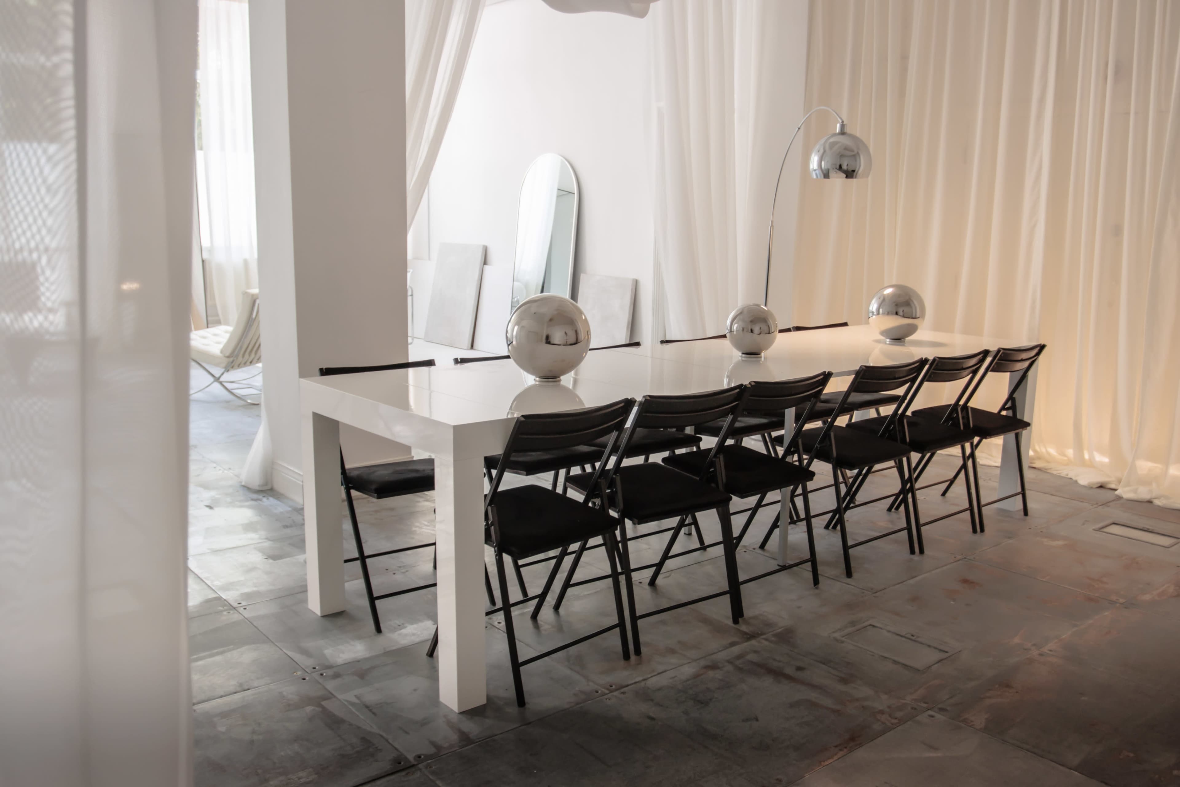A long white dining table with black chairs is set in a brightly lit room adorned with mirrors and sheer curtains.