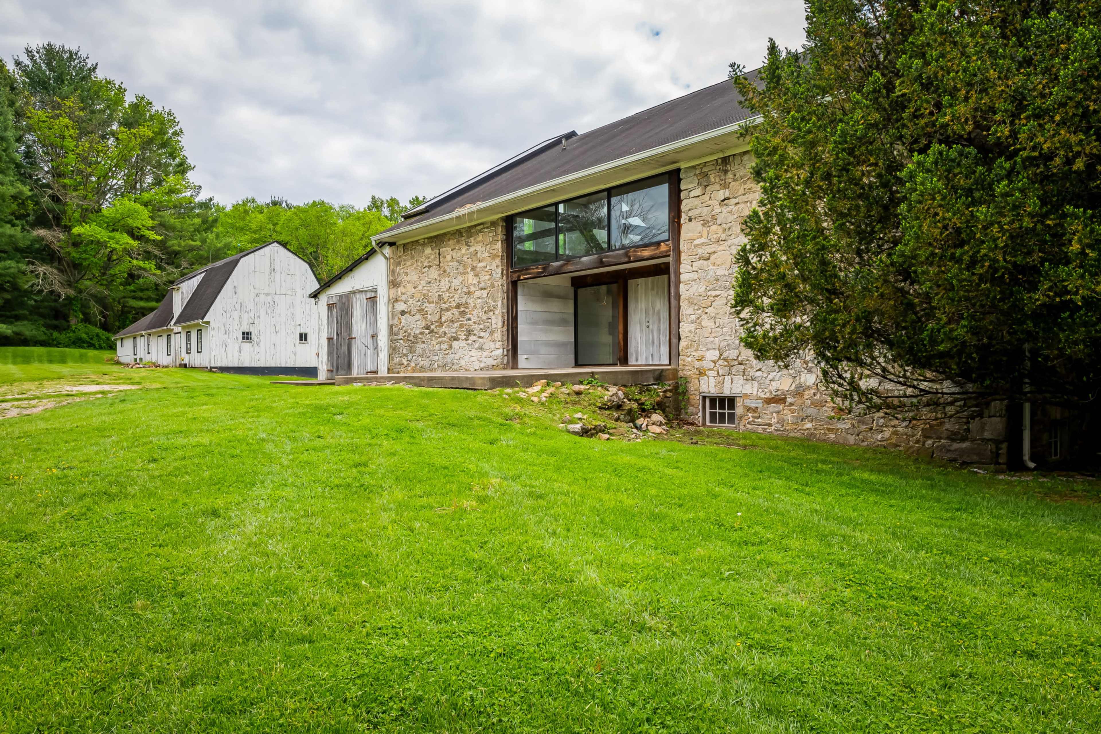 The image shows a stone barn with a modern design and large glass windows, accompanied by a white wooden barn in the background, all set on a grassy landscape.