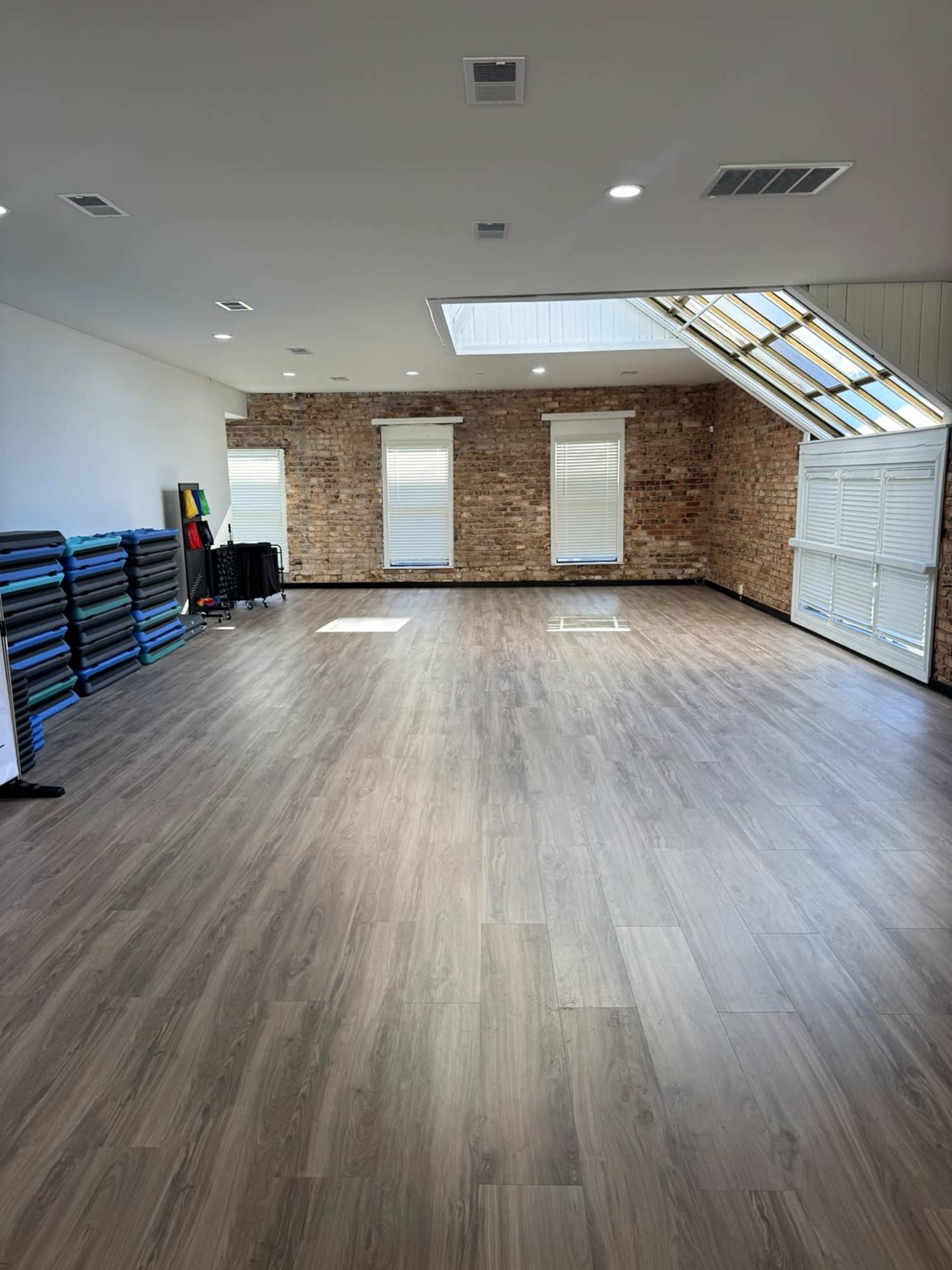 The image shows a spacious, empty room with wooden flooring, exposed brick walls, and large windows that allow natural light to enter.