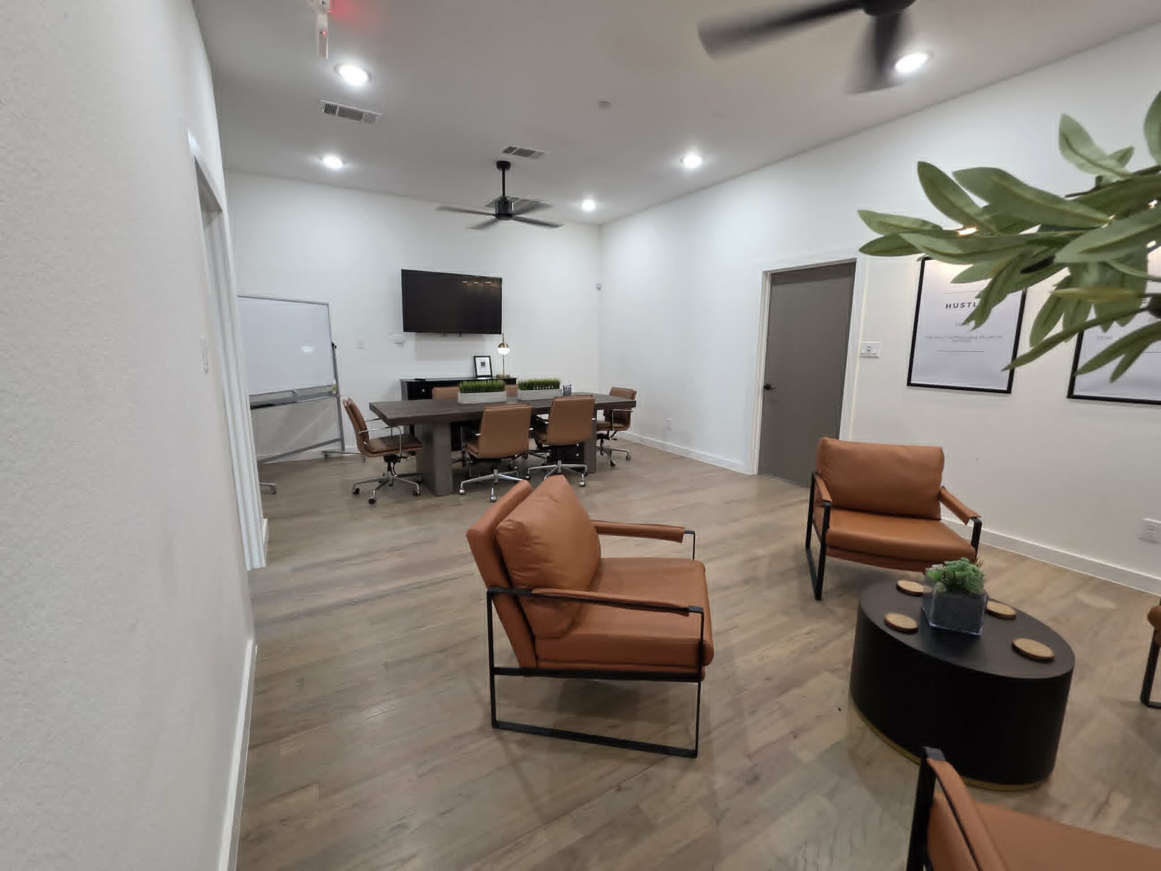 The image shows a modern office space featuring a conference table with chairs, a television on the wall, and a seating area with two armchairs and a coffee table.