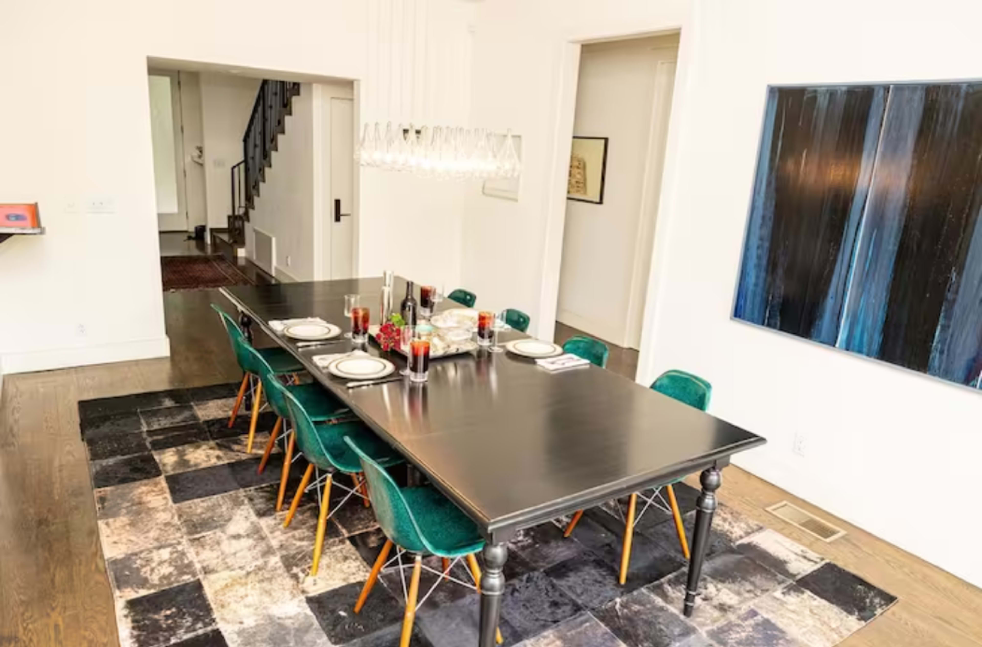 A dining room features a long black table surrounded by green chairs on a patterned rug, with a modern light fixture overhead.