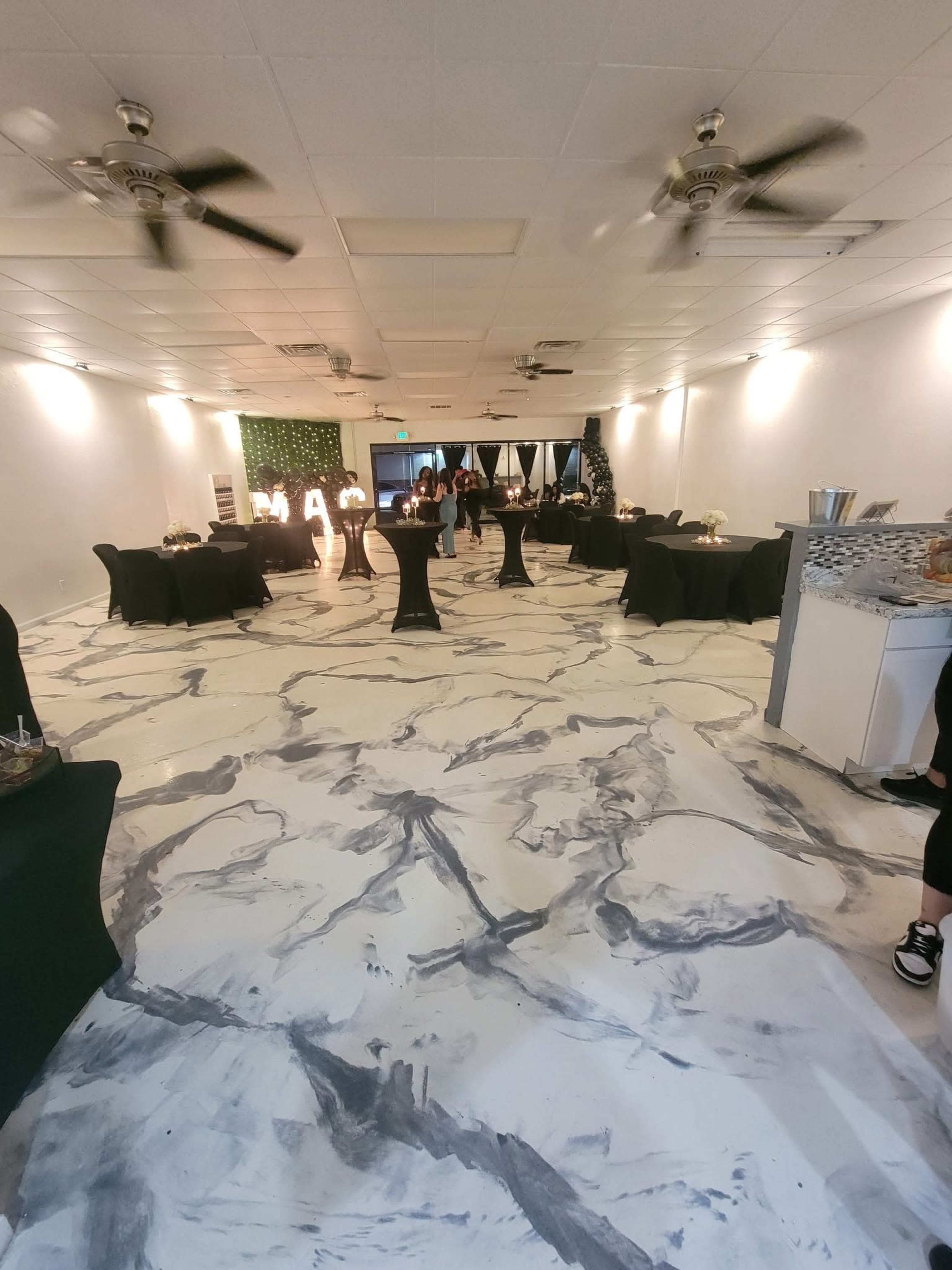 The image shows a spacious event room with black tables and chairs, a marbled floor, and low lighting, where guests are gathered around a presentation area.