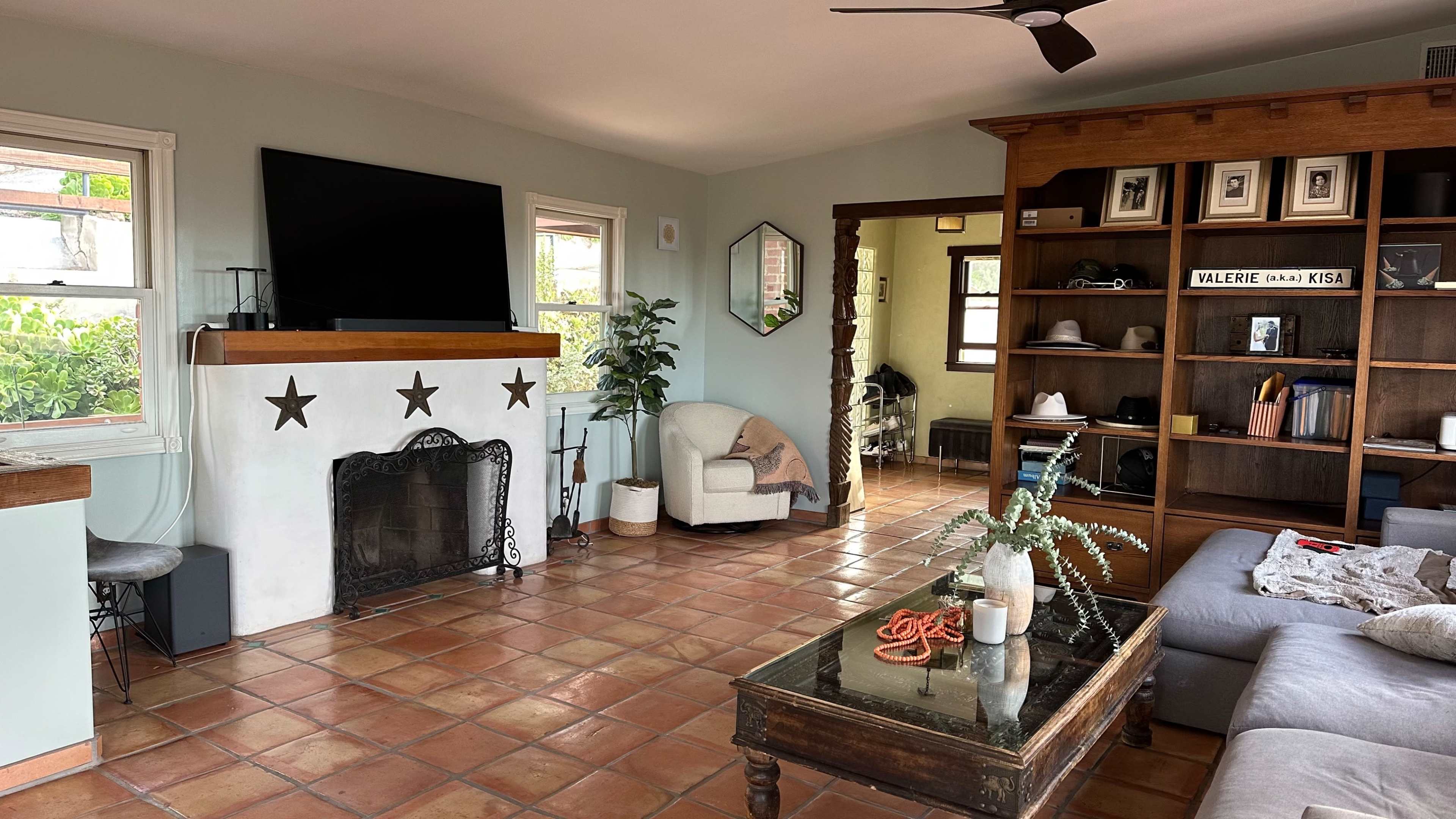 A spacious living room with a fireplace, a large TV mounted on the wall, a gray couch, and a wooden shelving unit filled with decorative items.