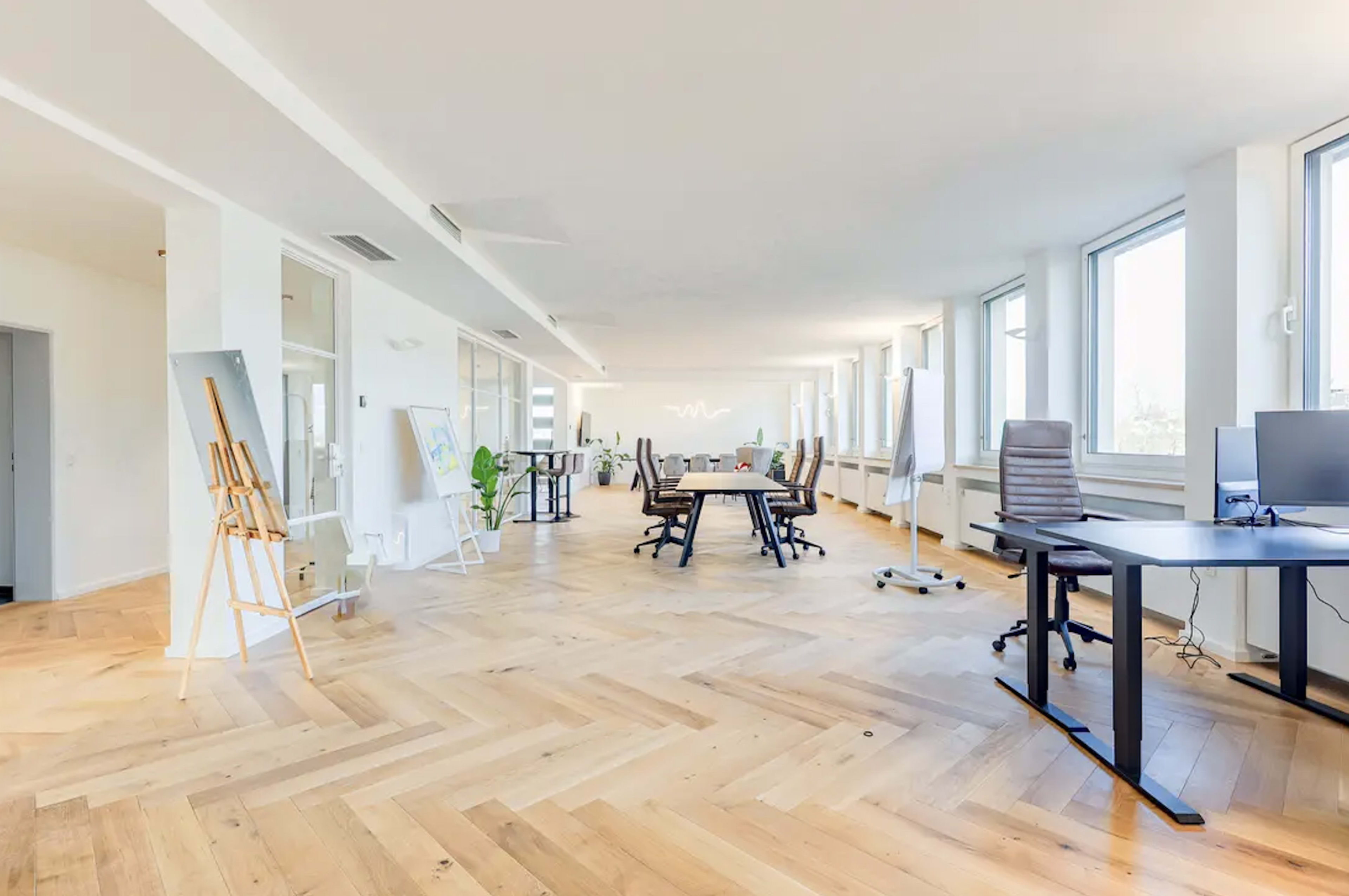 The image depicts a spacious, well-lit office featuring wooden flooring, a central conference table surrounded by chairs, and workstations with desks and computers near large windows.