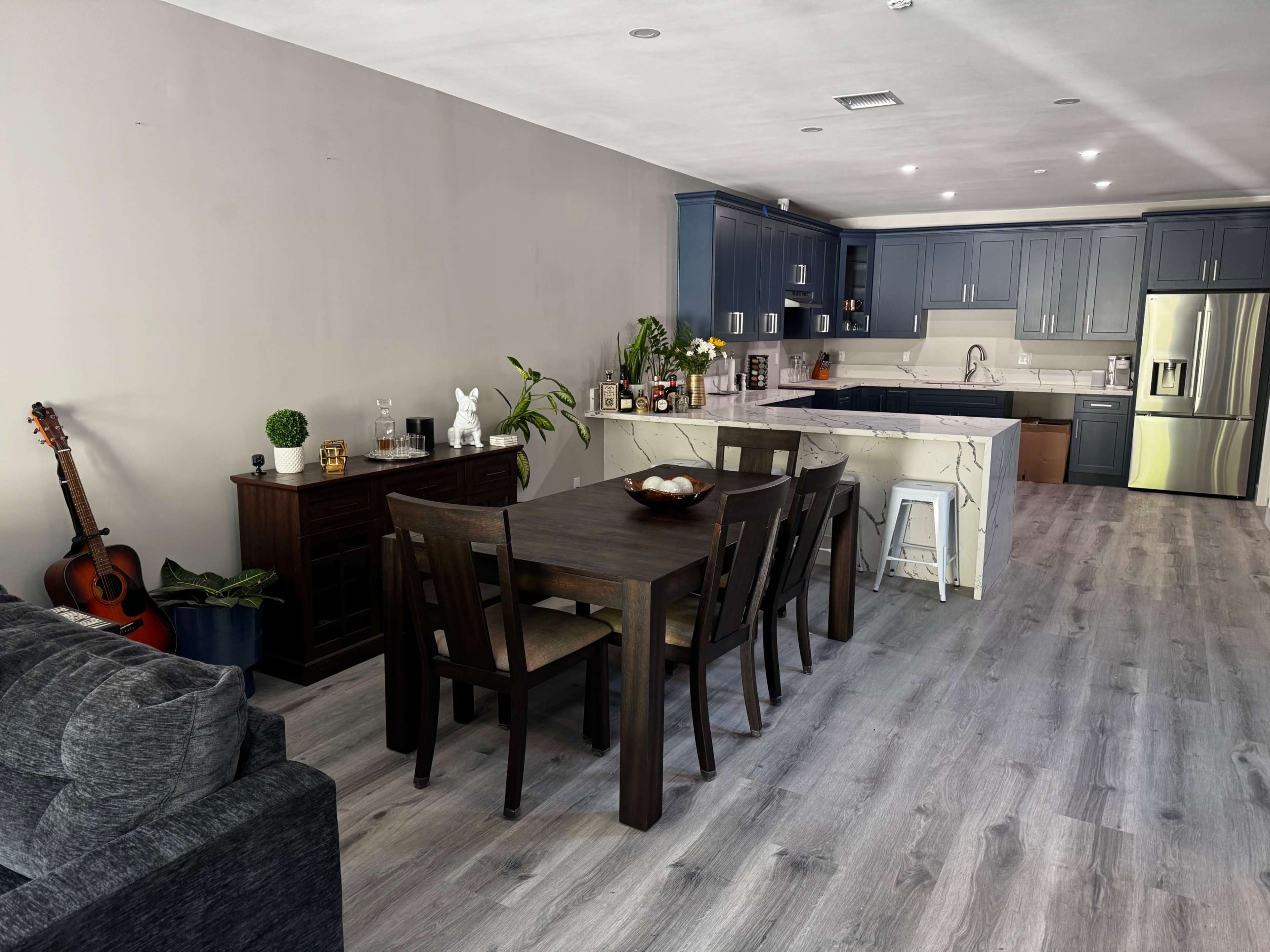 A modern open-concept living space features a dining area with a dark wooden table and chairs, a kitchen with navy cabinets, and a guitar leaning against the wall.
