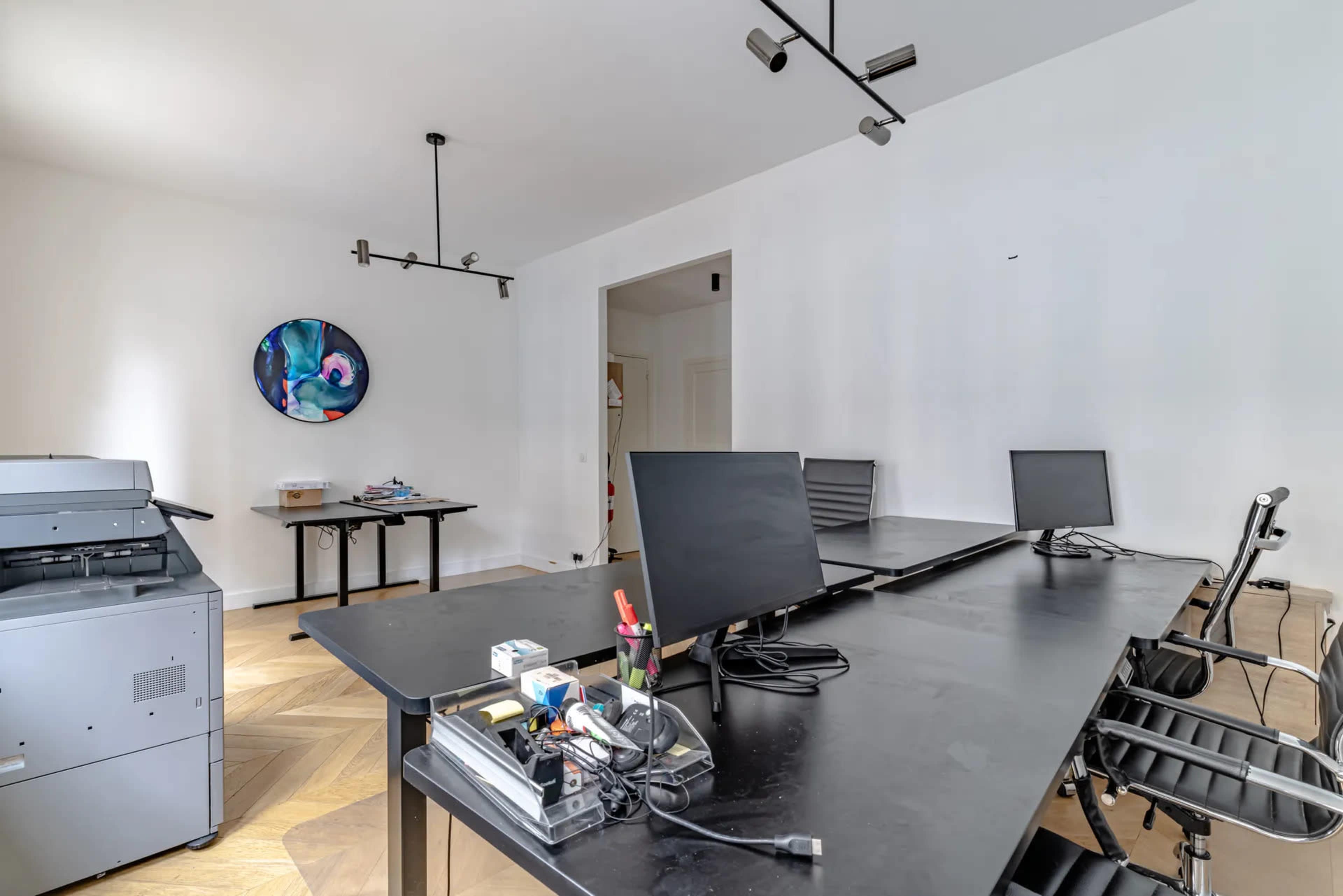 The image depicts a minimalistic office space featuring two desks, a printer, and two computer monitors on a black tabletop.