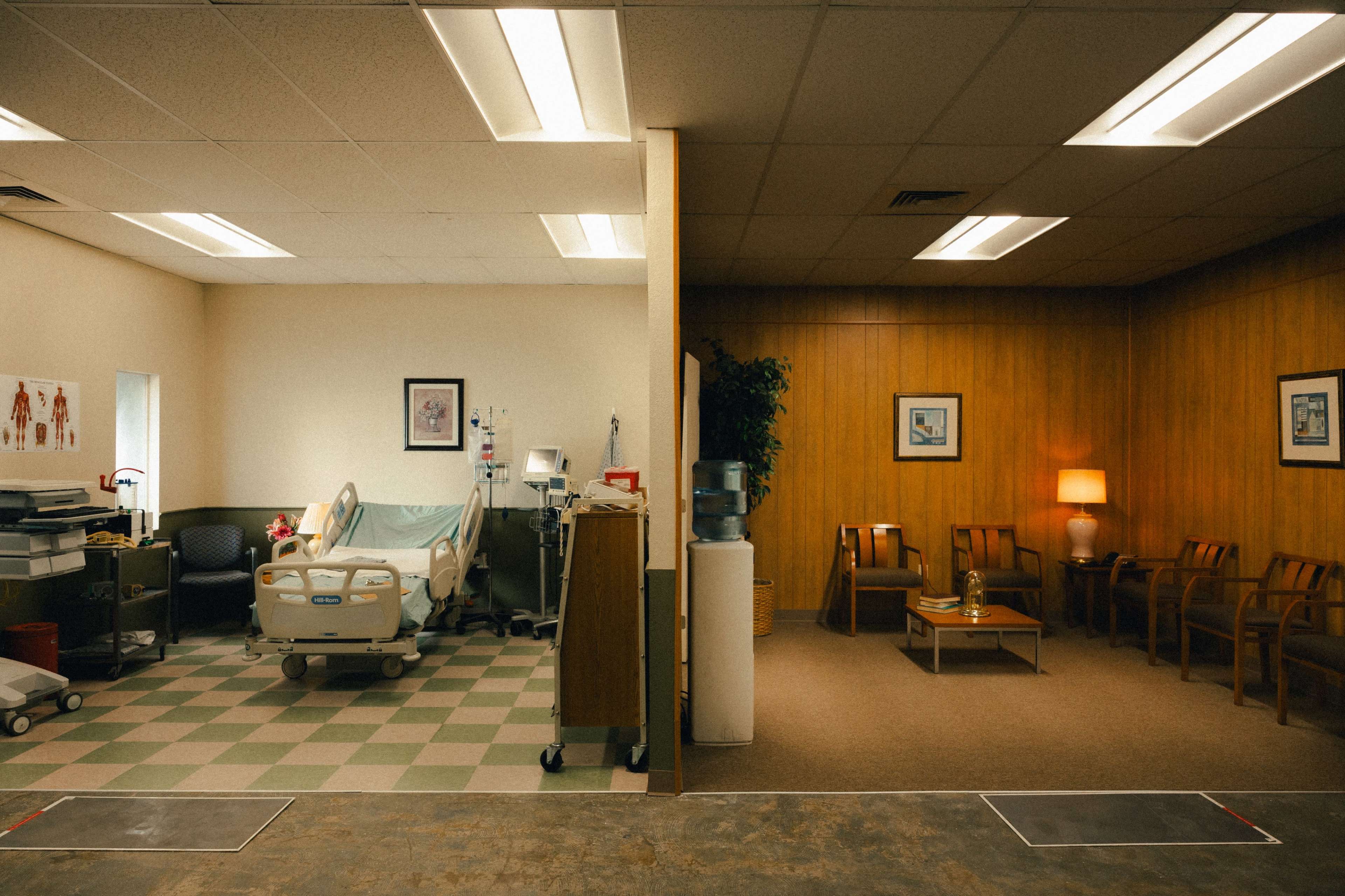 A split room, with a hospital bed and medical equipment on one side and a waiting area with chairs and a table on the other.