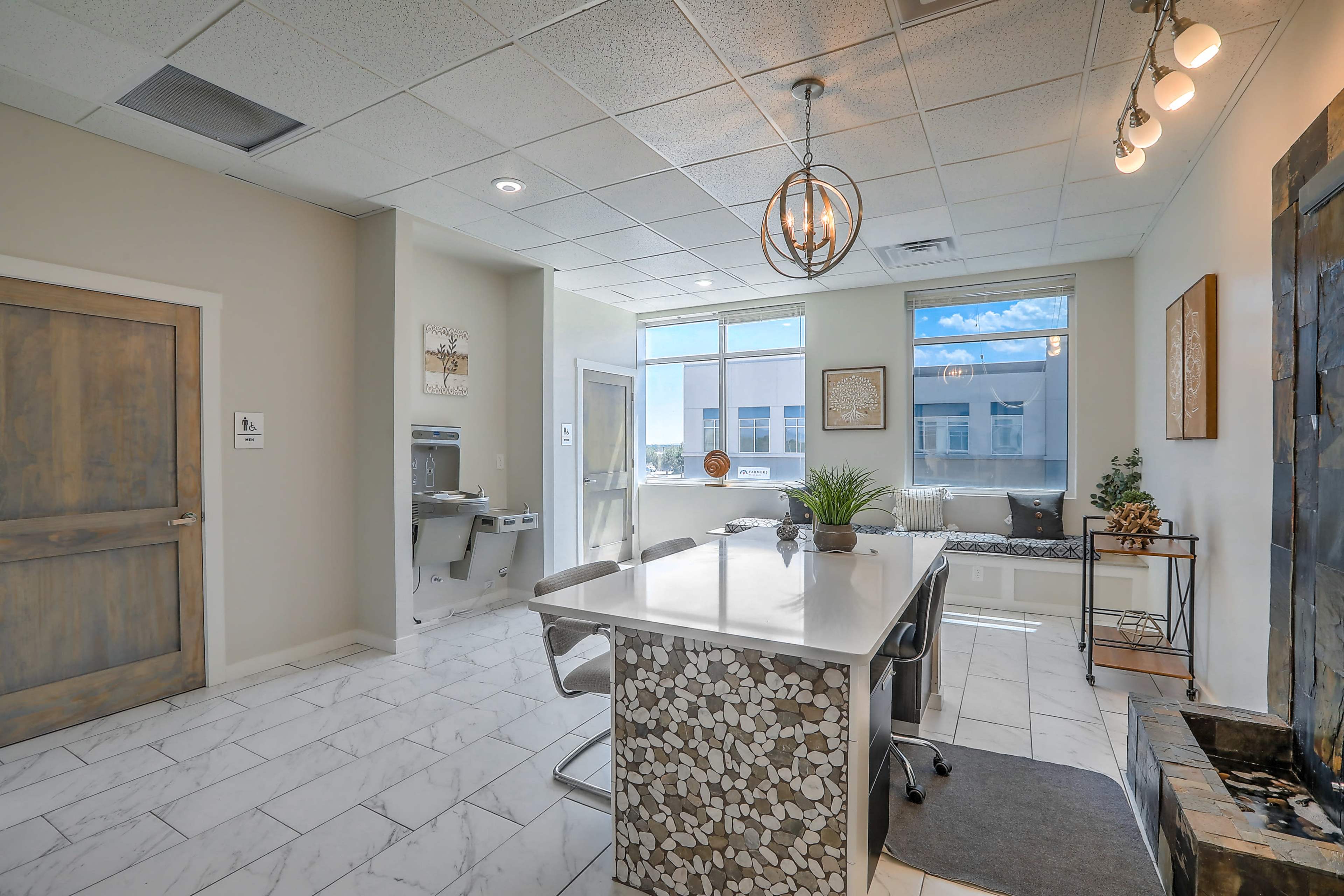 A modern office space featuring a large island desk with a mosaic design, windows allowing natural light, and a minimalist decor.