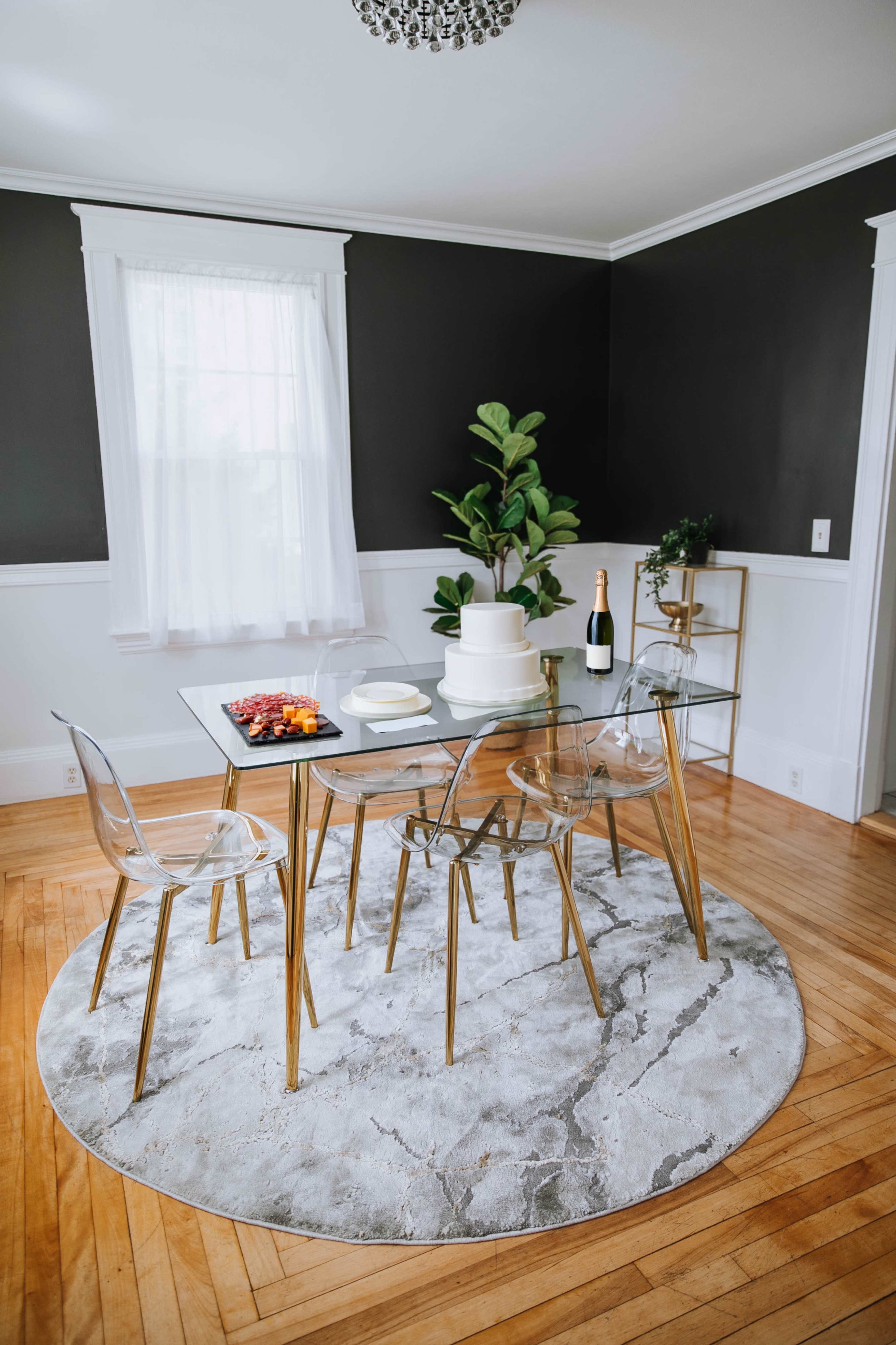 A glass dining table is set with plates and a fruit platter, surrounded by clear chairs on a circular rug in a room with dark walls and a potted plant.