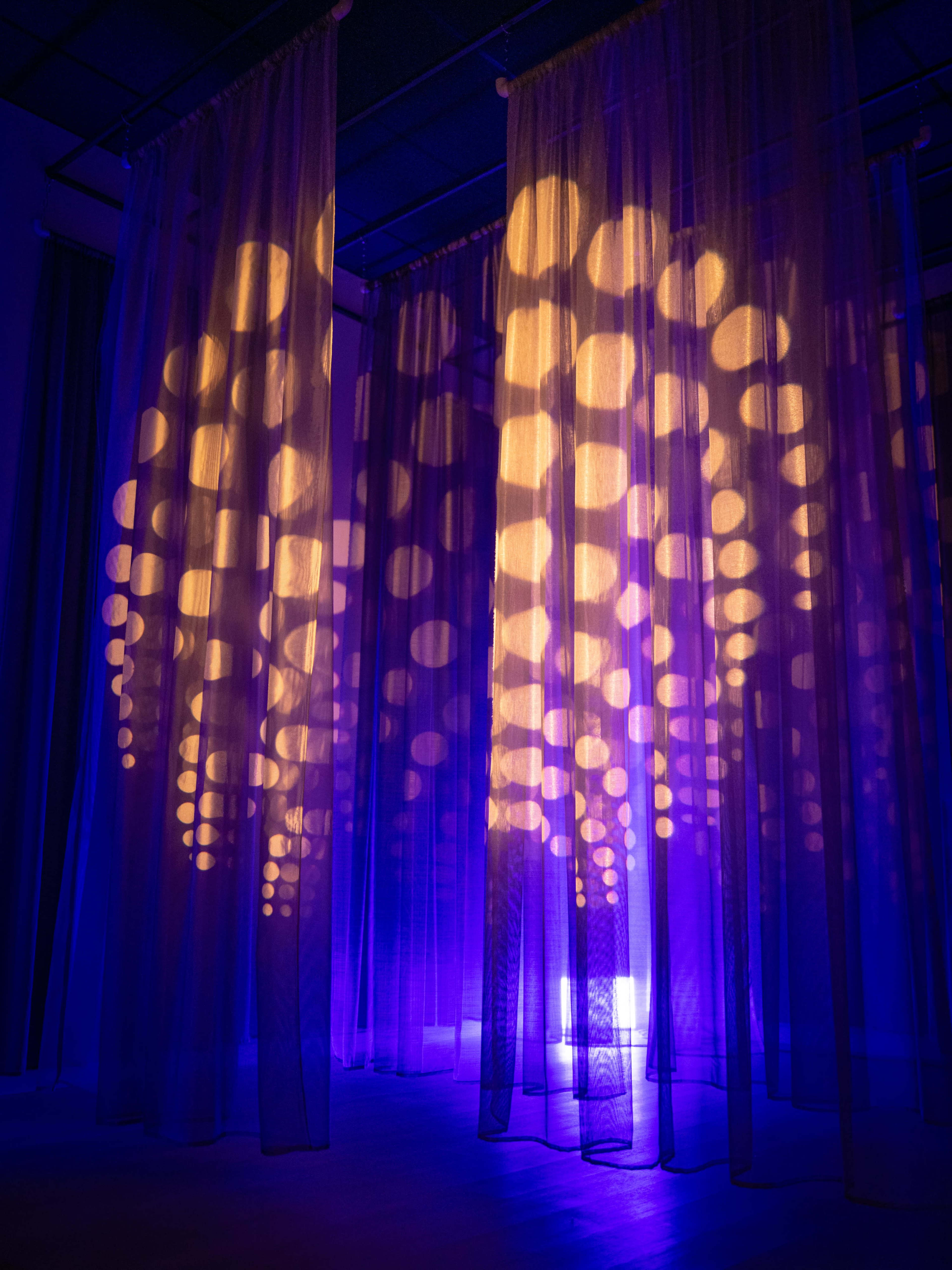 Translucent curtains hang in a dimly lit space, casting circular light patterns in shades of purple and orange.