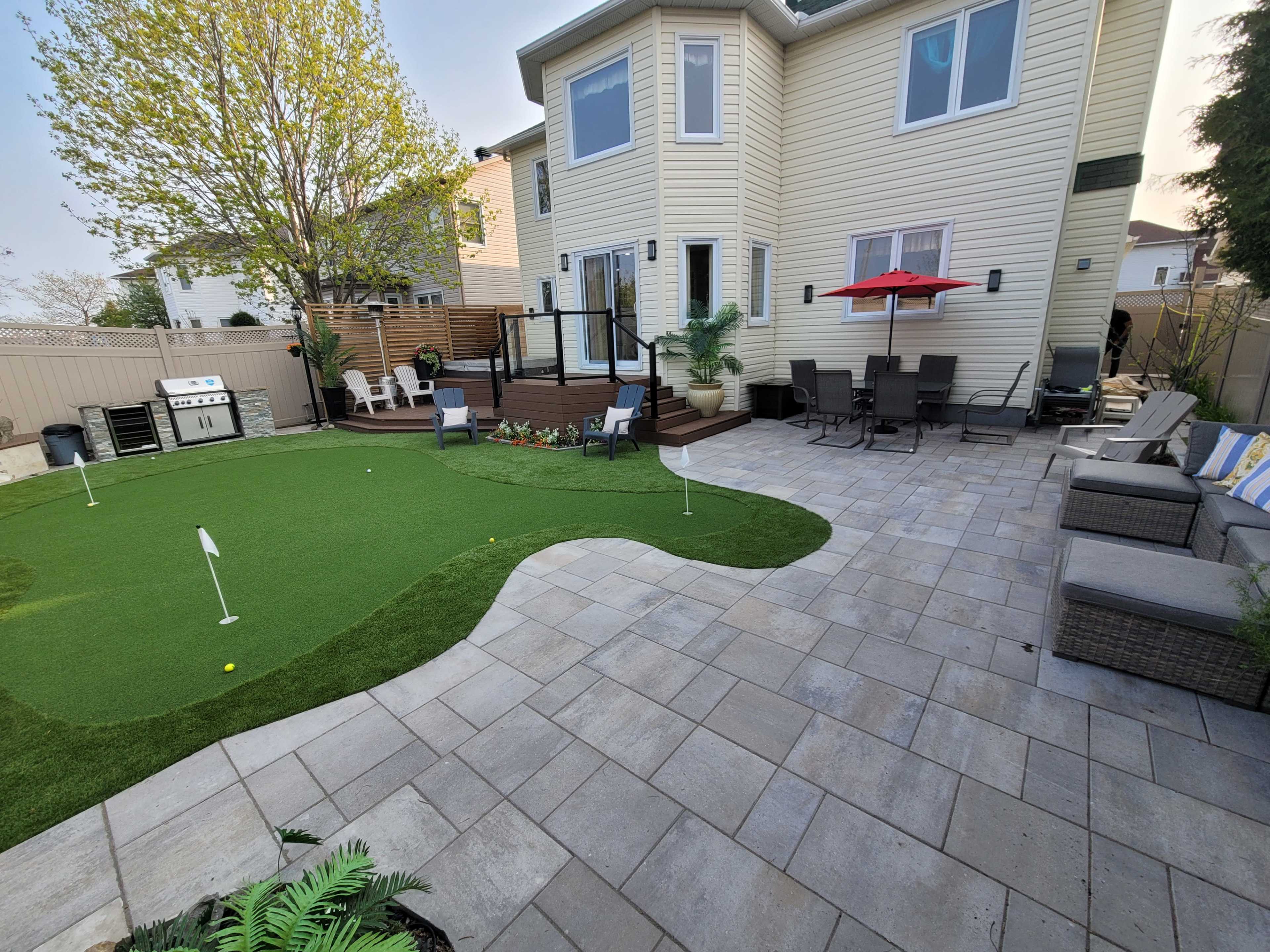 A landscaped backyard featuring a patio with seating, a putting green, and a house with a deck.