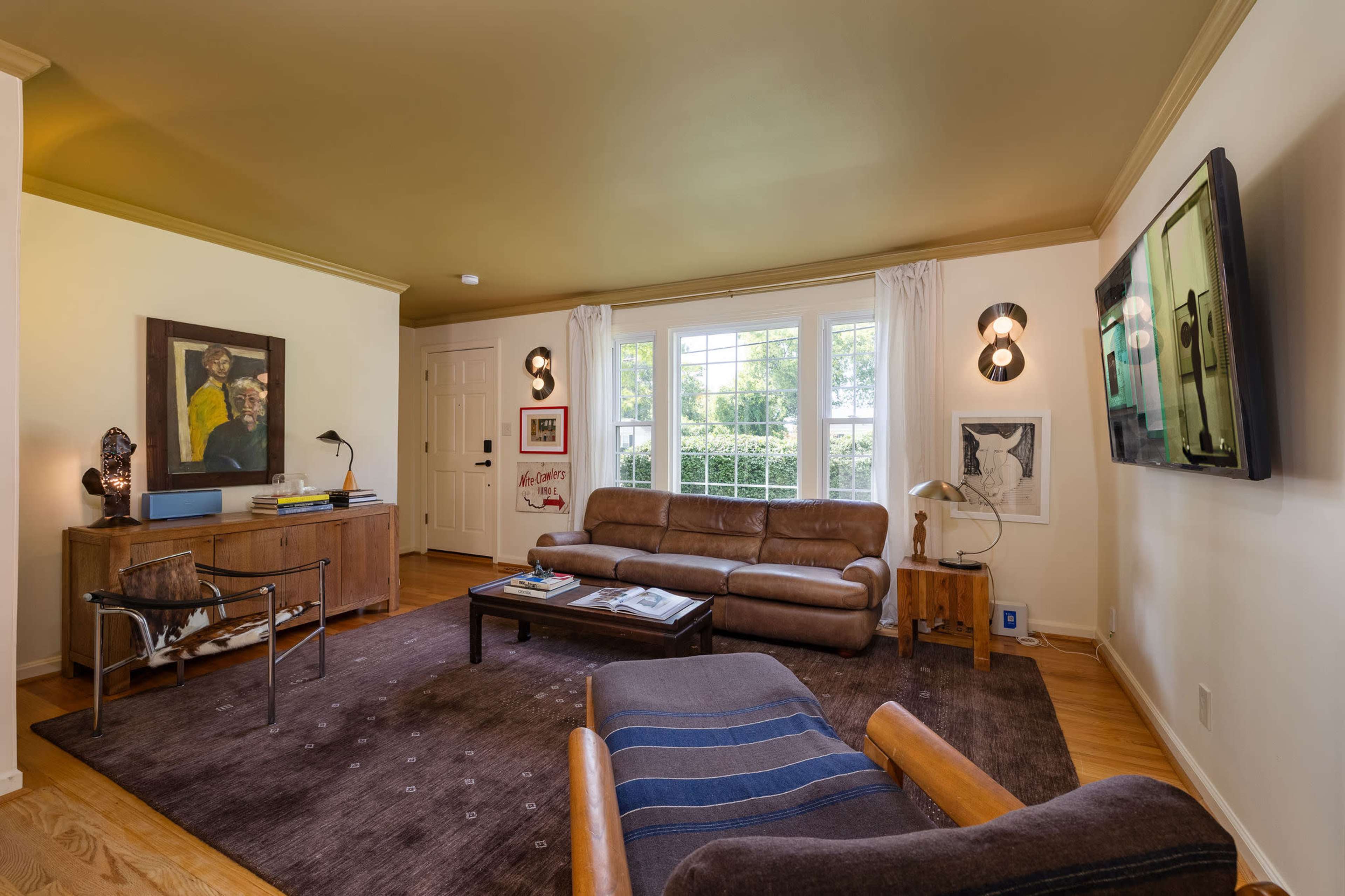 The living room features a brown leather sofa, a wooden coffee table, and two windows with white curtains, illuminated by wall-mounted lights.