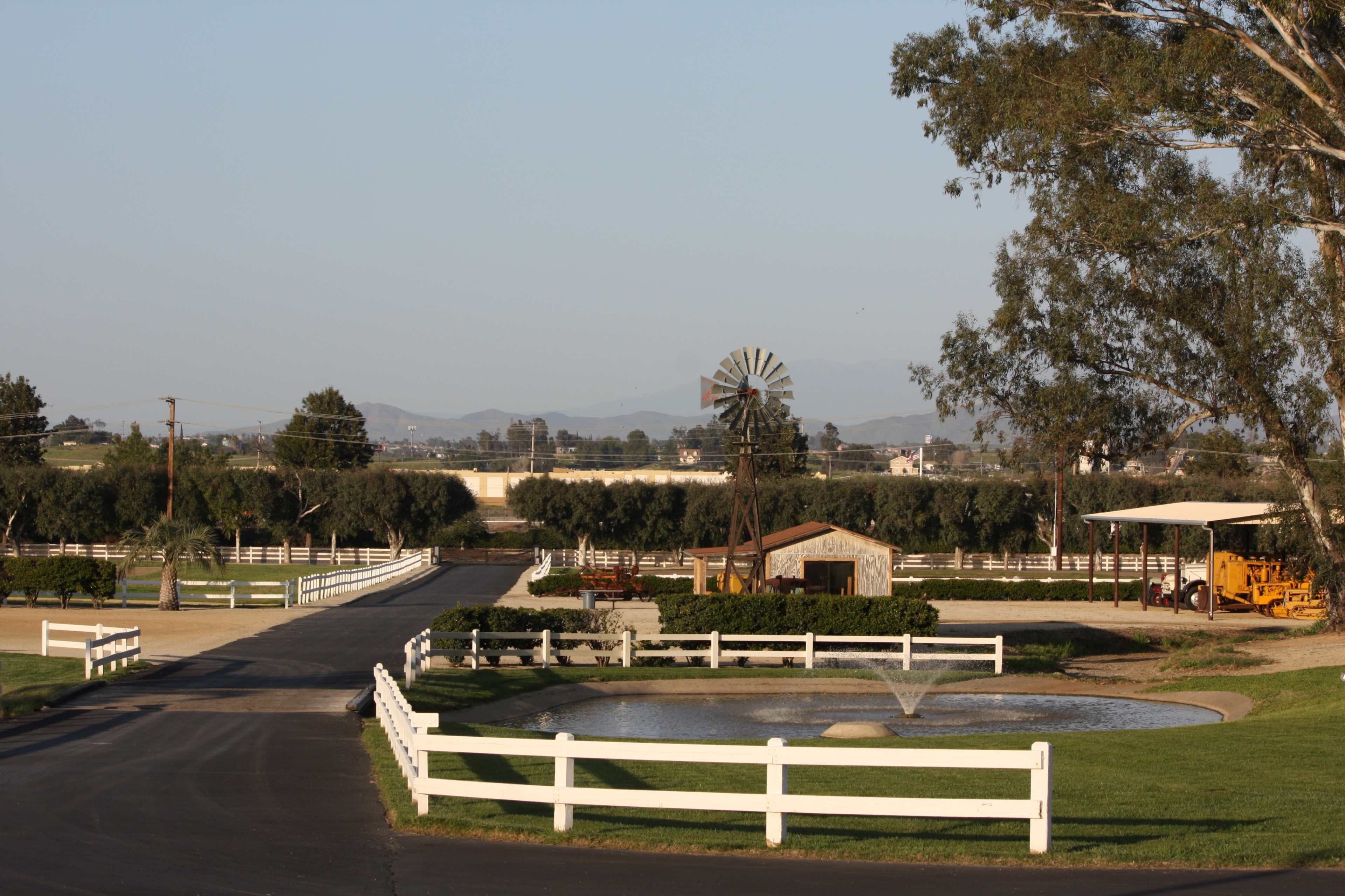 A ranch landscape with a driveway leading to a small pond, a windmill in the background, and fenced areas with trees and buildings.