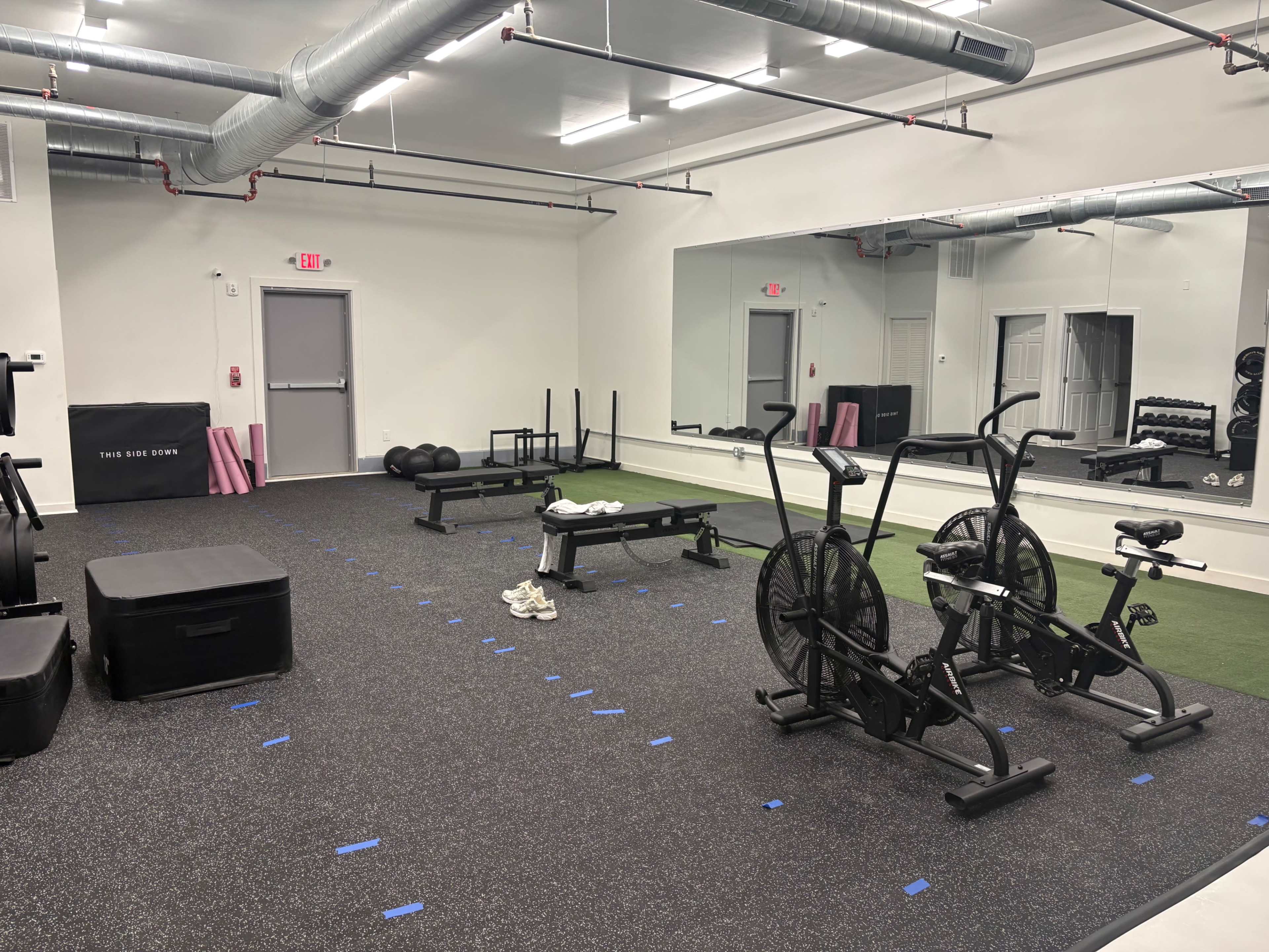 The image shows a gym space with various exercise equipment, including an exercise bike, a weight bench, and a turf area, all set on a rubberized floor.