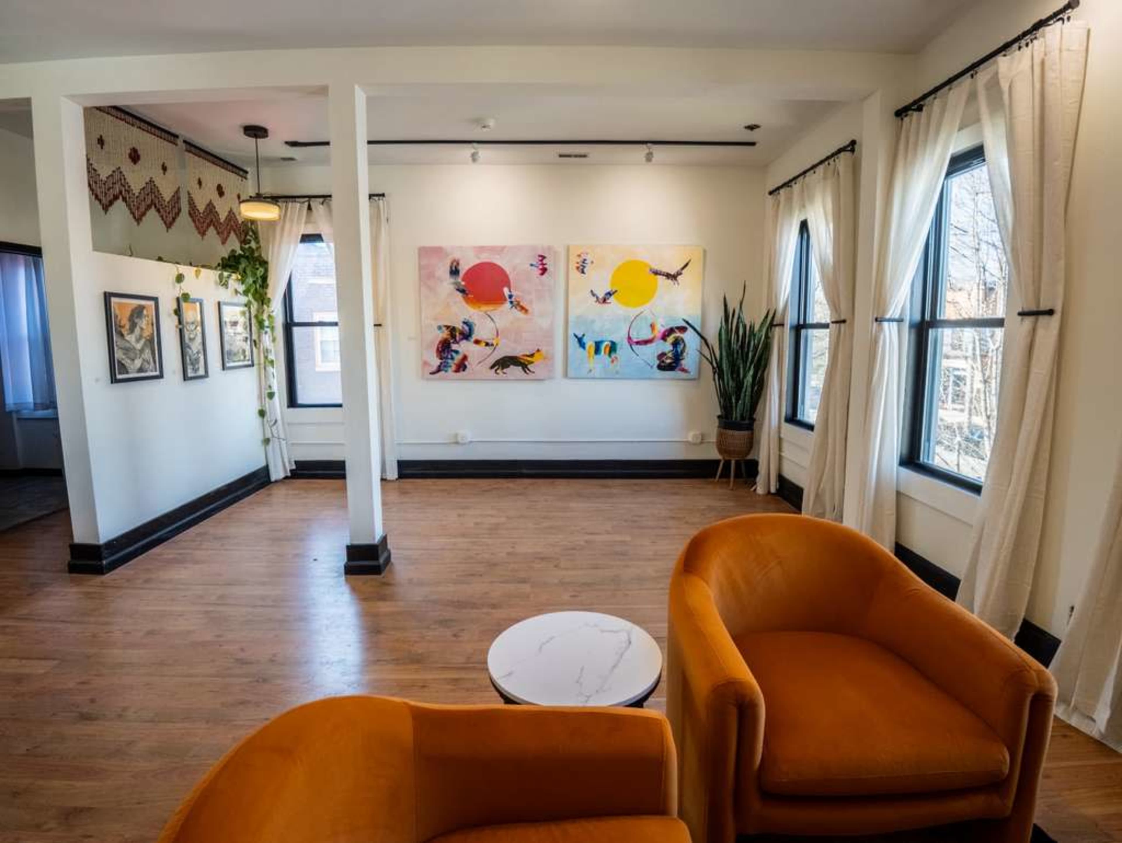 The image shows a bright, spacious room with wooden floors, featuring two orange chairs and large windows, along with colorful abstract artwork on the wall.