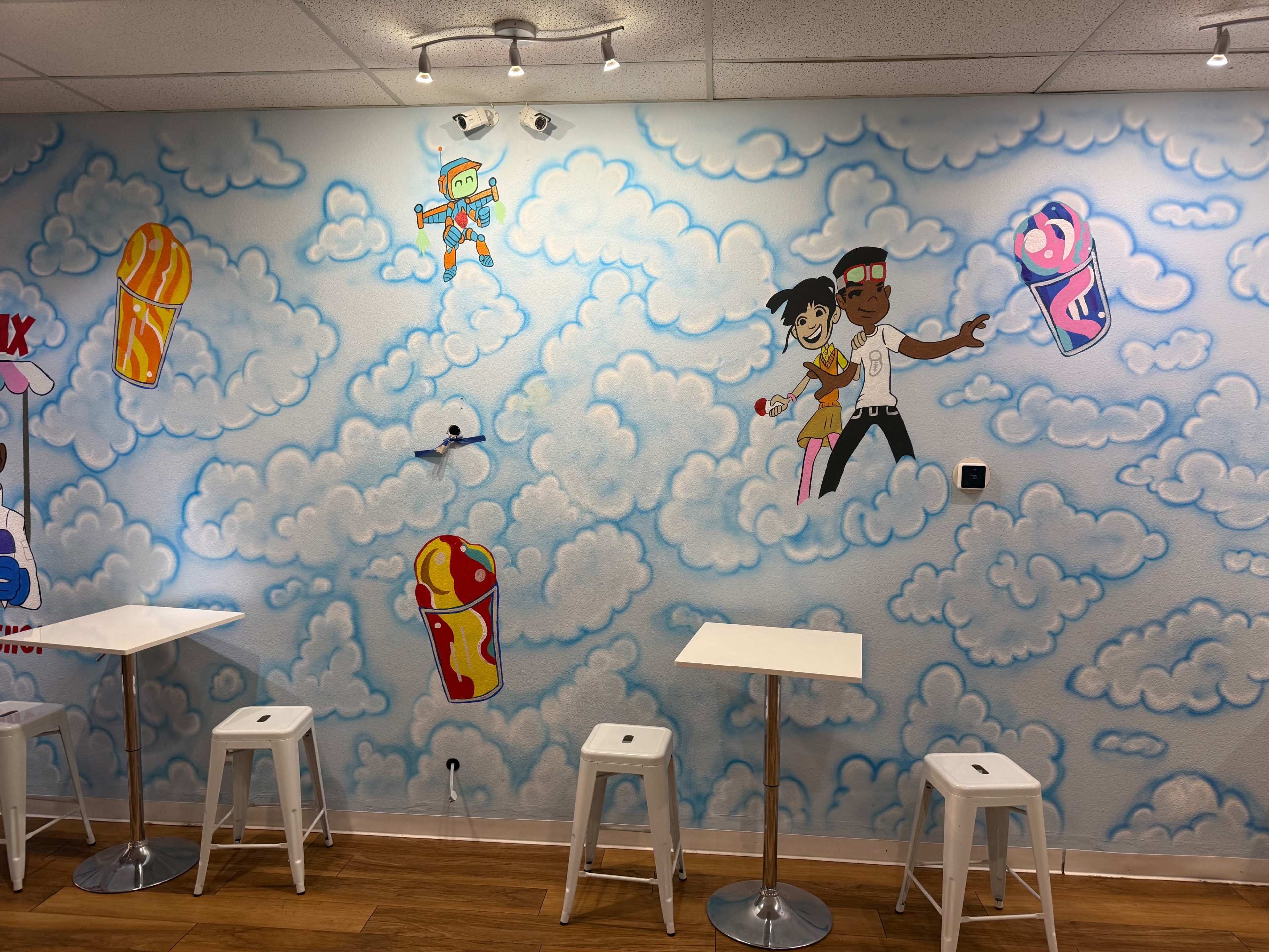 The image shows a wall mural featuring cartoon characters and colorful ice cream cones set against a backdrop of fluffy clouds, with white tables and stools in the foreground.