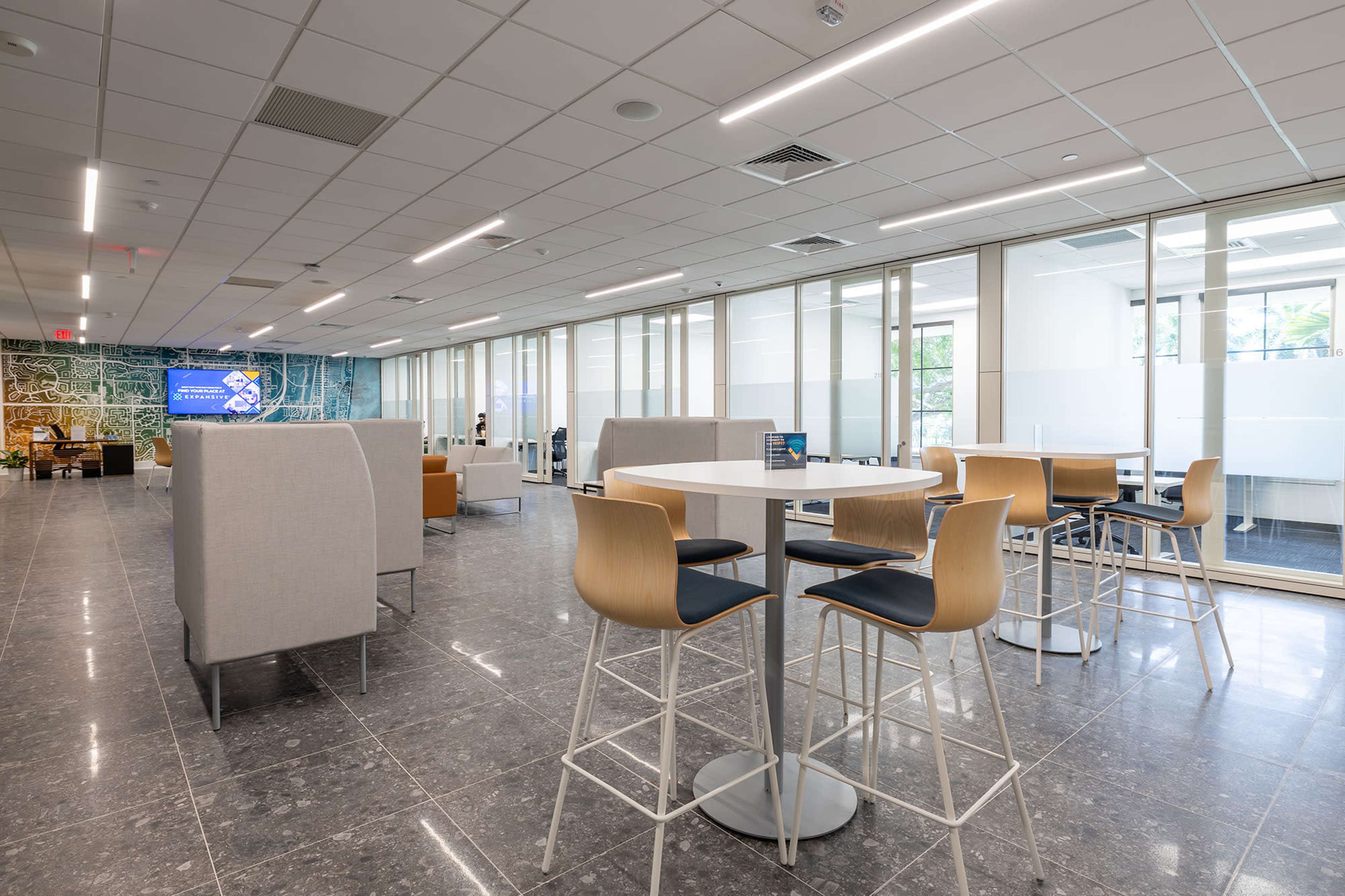 The image shows a modern, open-plan office space with several high tables and stools, along with partitioned seating areas and large glass windows allowing natural light.