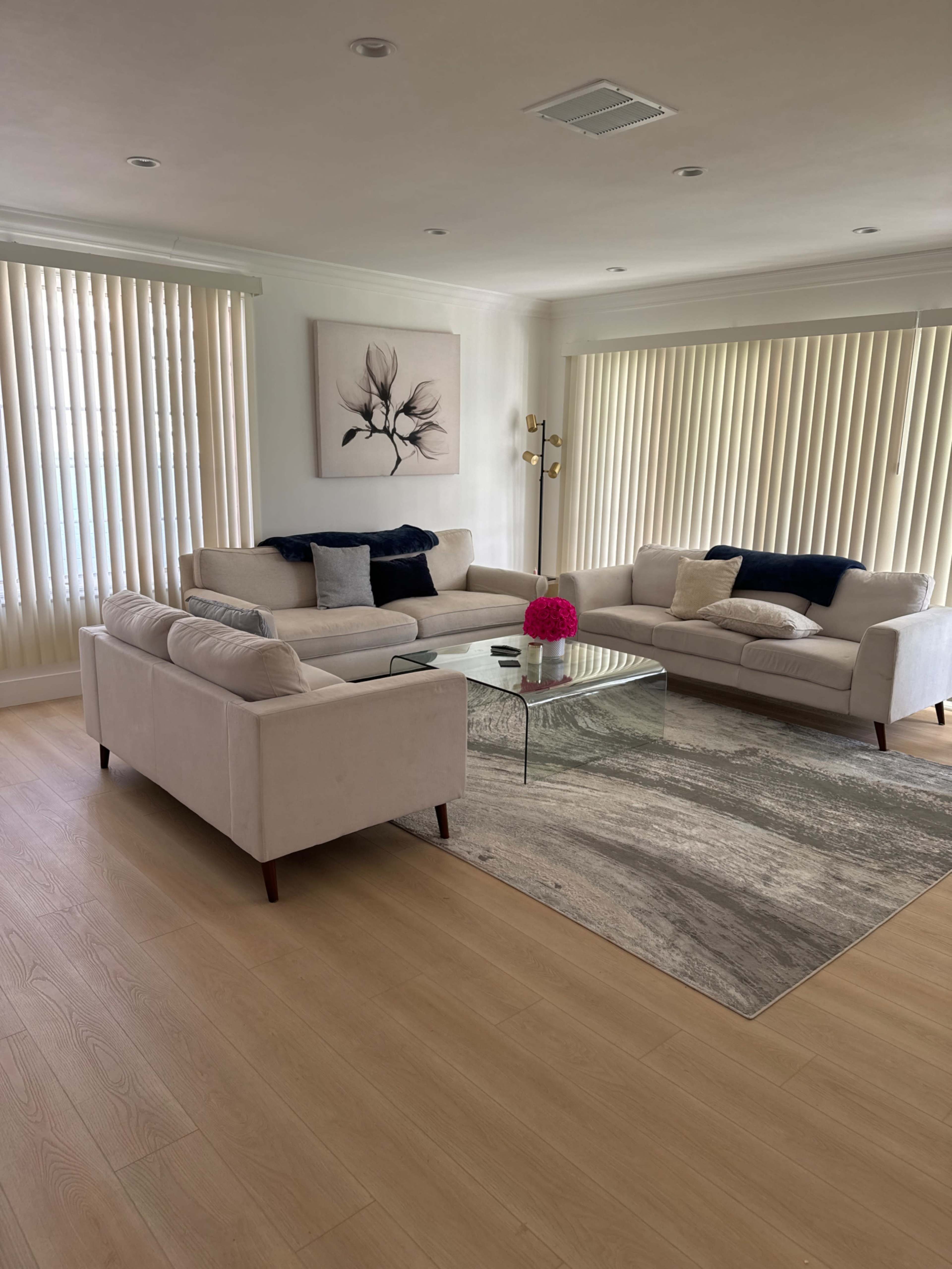A spacious living room features a light-colored sectional sofa, a glass coffee table, and large windows with vertical blinds.