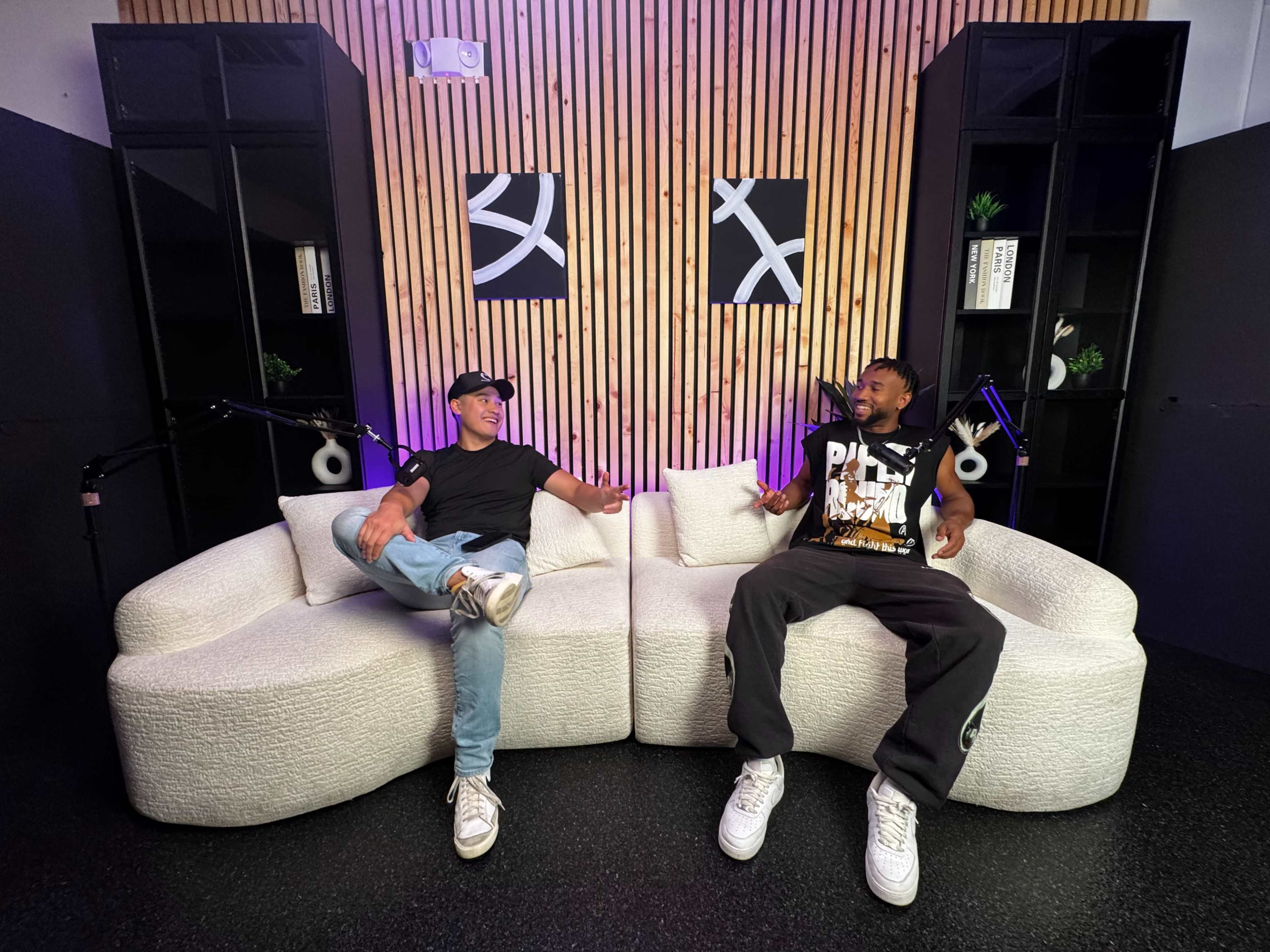 Two men sit on a light-colored sofa in a modern studio with wooden walls and decorative shelves, engaged in conversation.