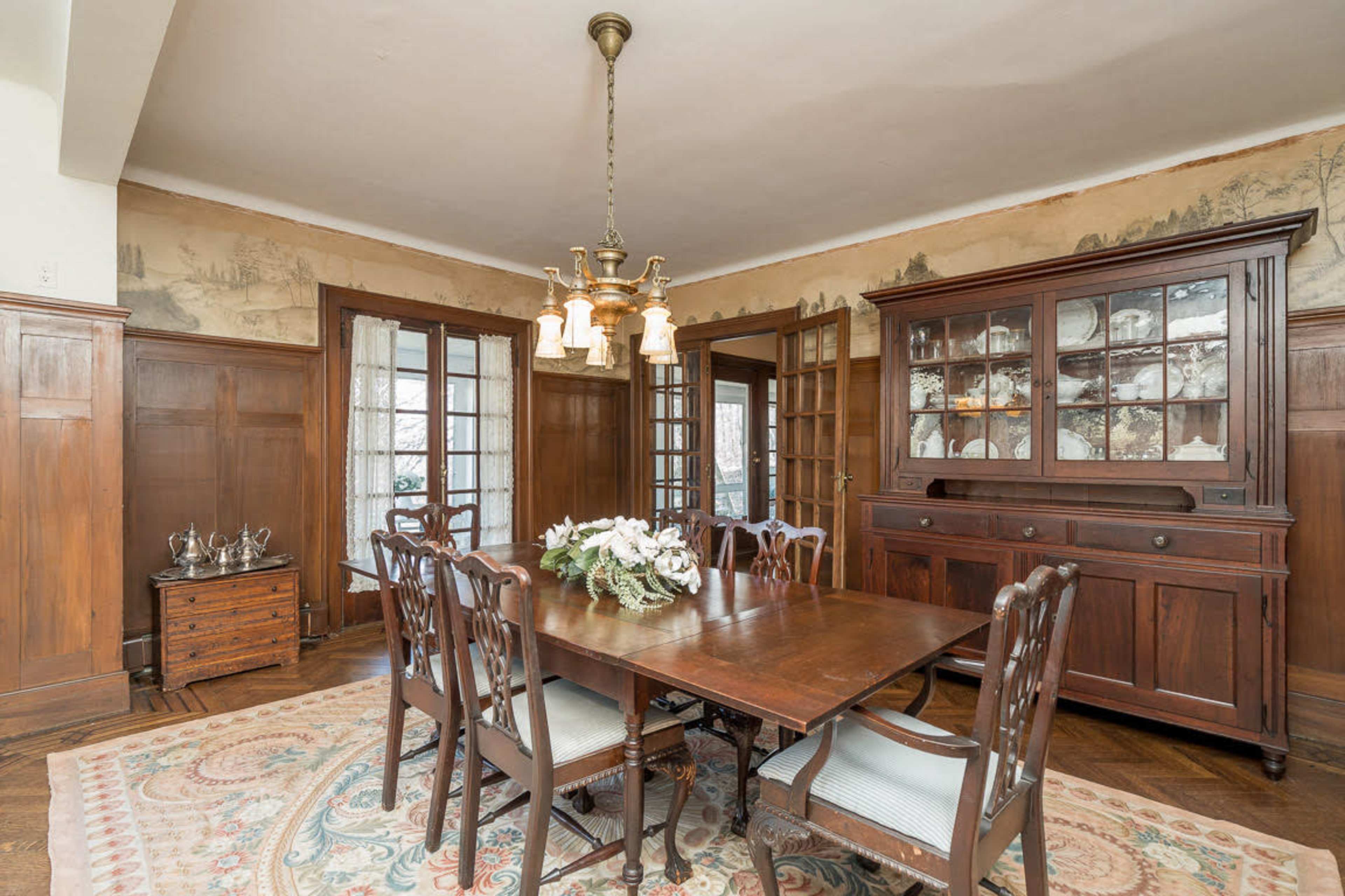 A dining room features a large wooden table surrounded by chairs, with a vintage china cabinet and a chandelier above.