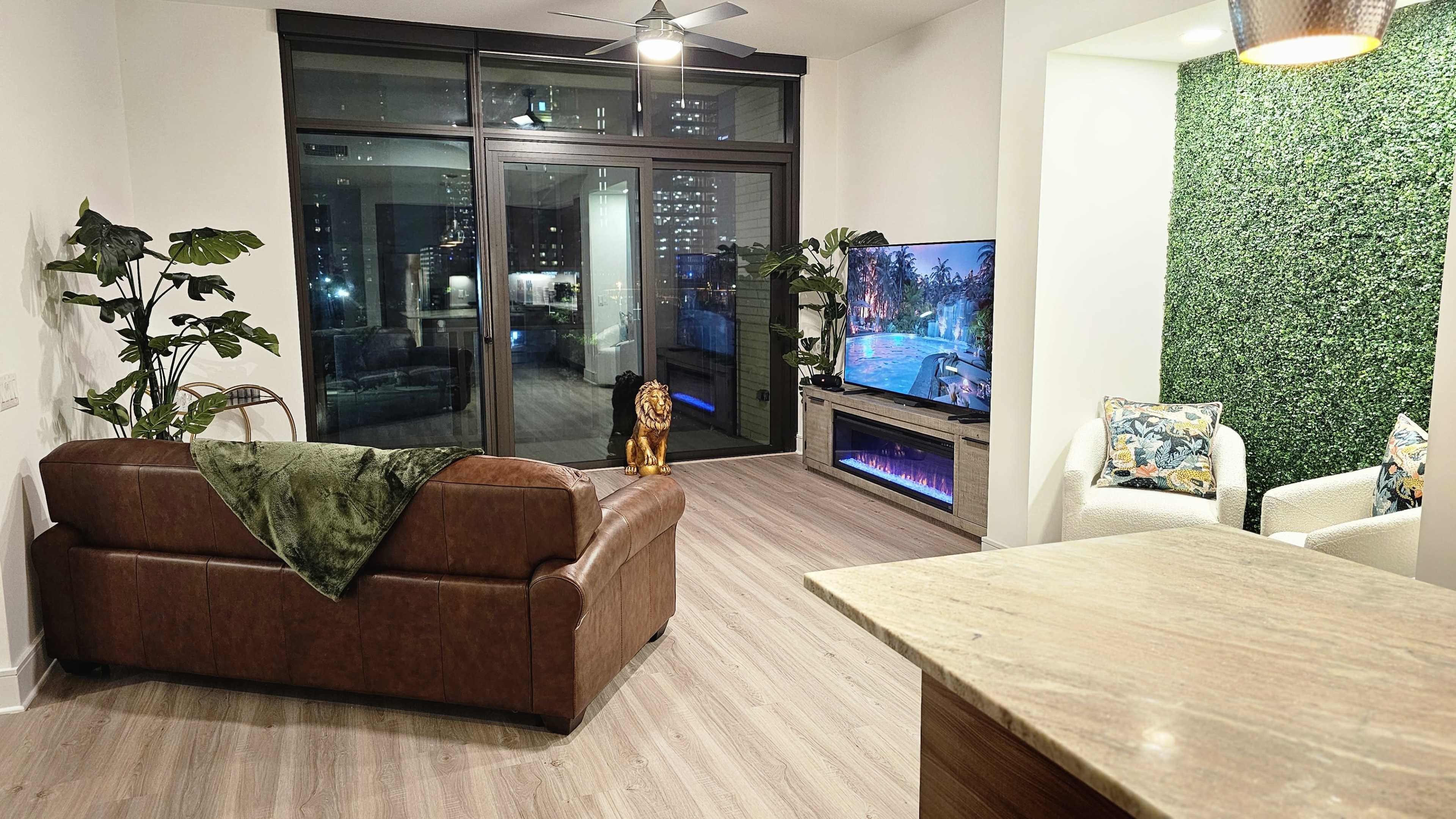 A modern living room features a brown leather sofa, a large television displaying a pool scene, and greenery on the walls, with a view of a city skyline through glass doors.
