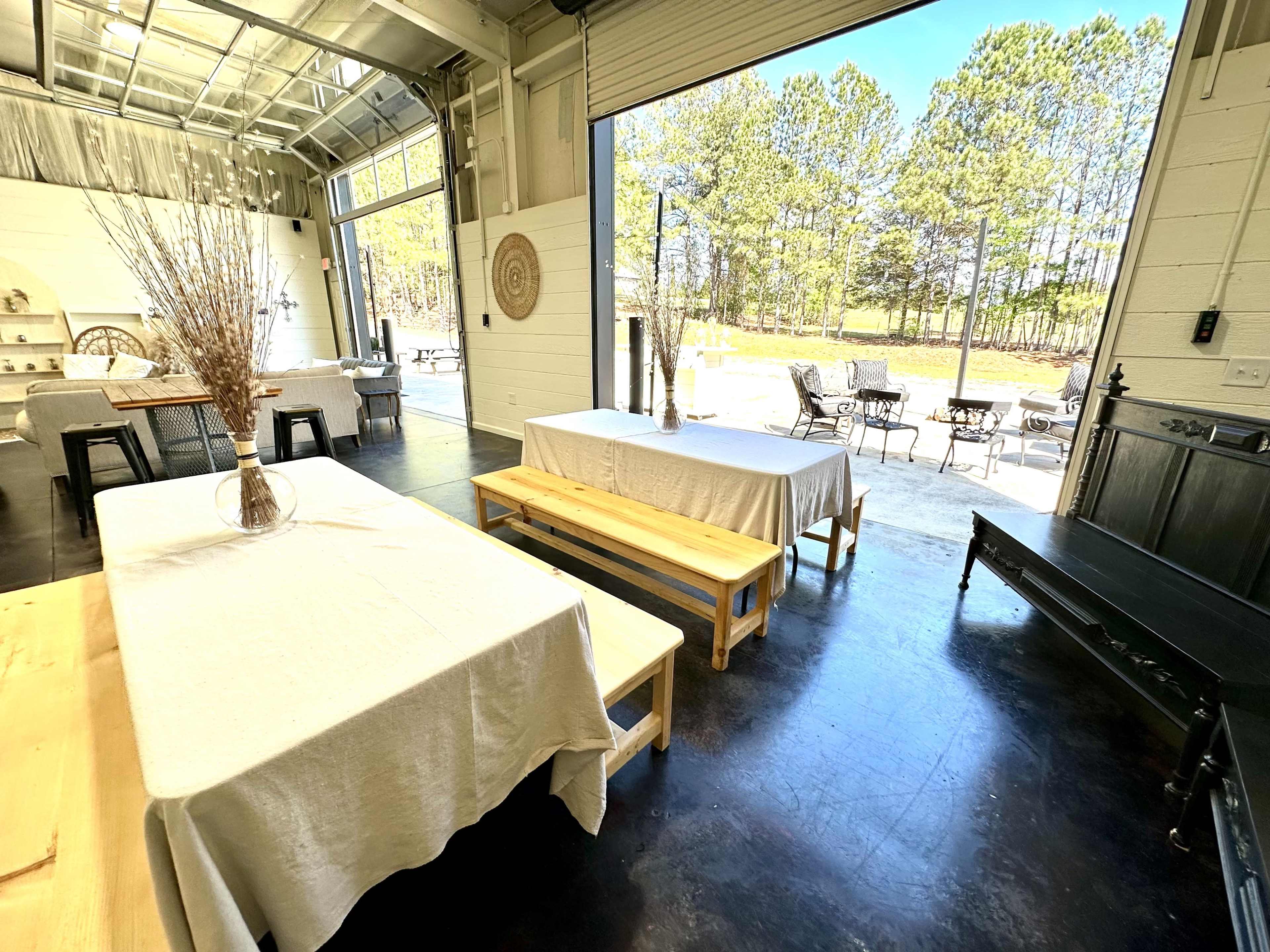 A spacious indoor area with tables covered in white linens, leading to an open door that reveals a view of a tree-lined outdoor space.