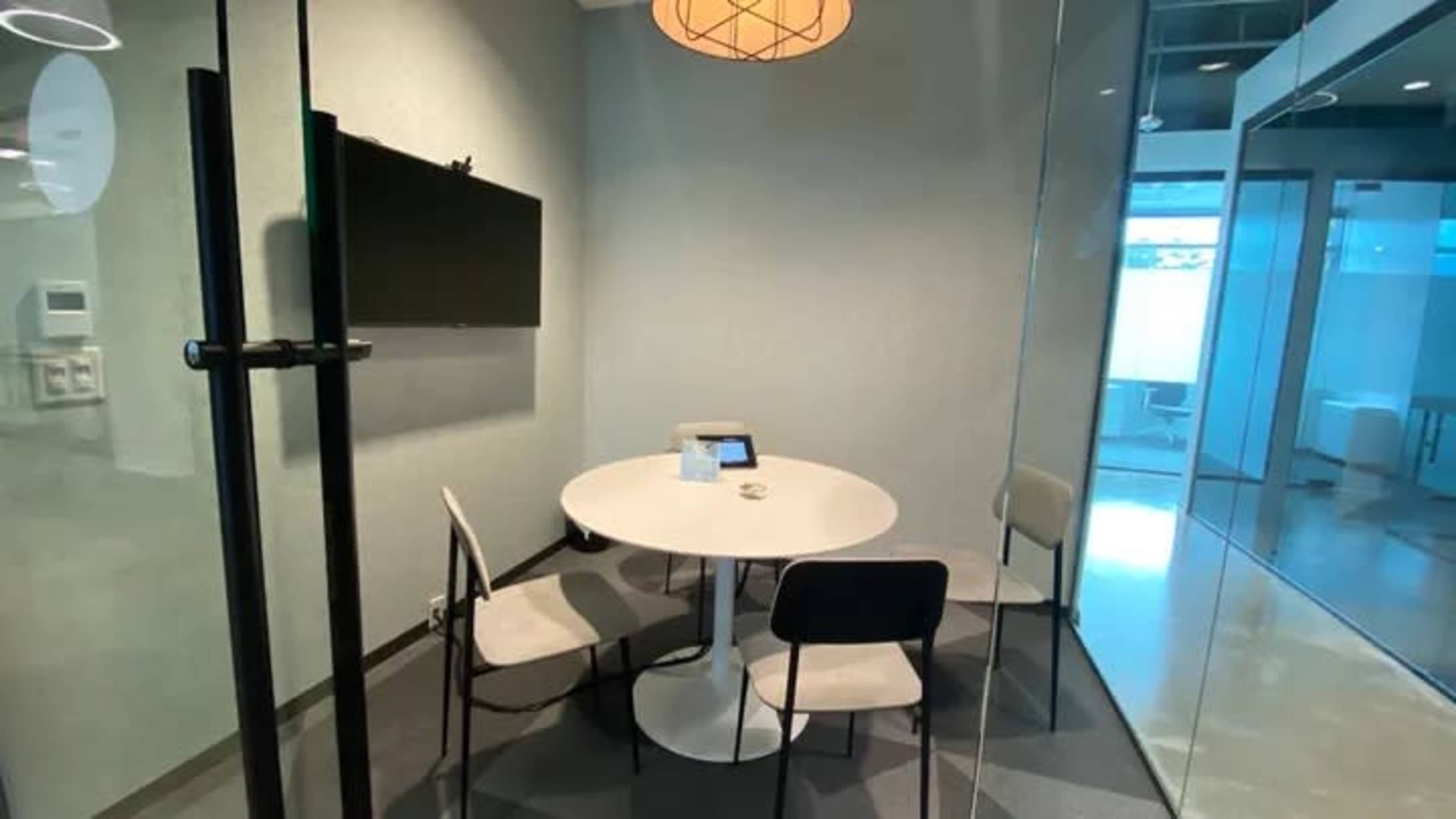 A conference room with a round white table, four chairs, and a wall-mounted TV, featuring glass walls and a pendant light.